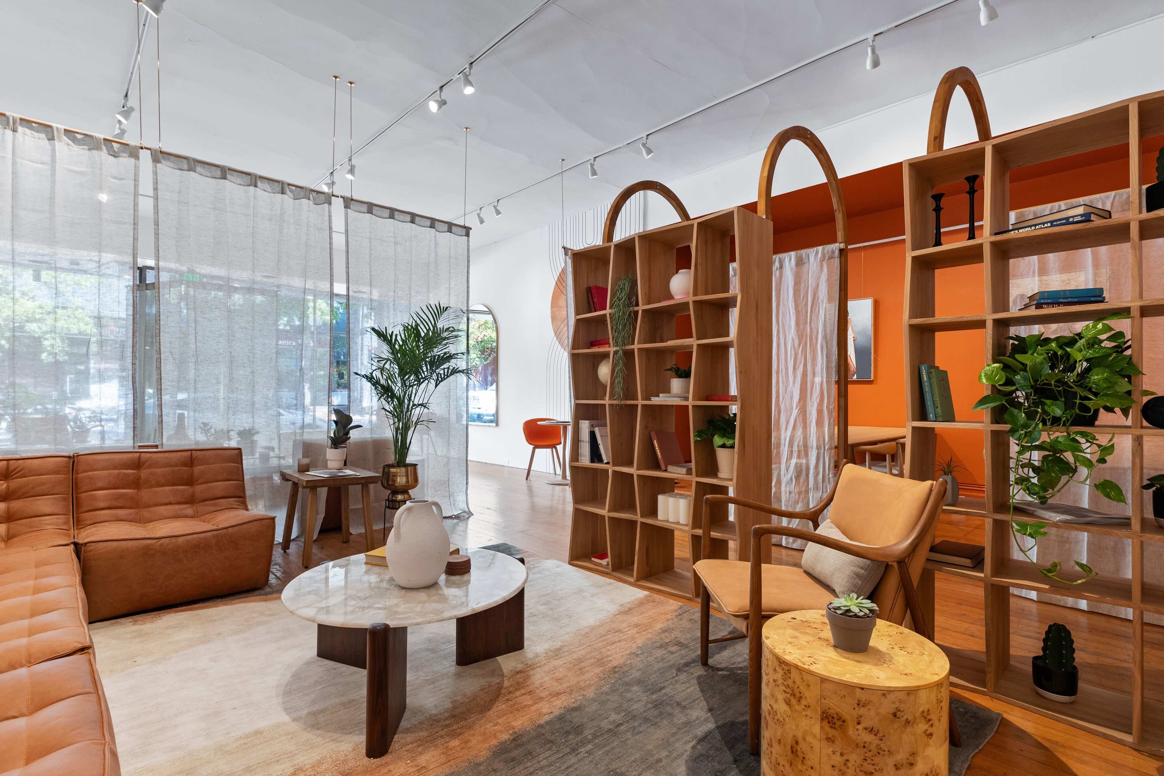 A modern living space featuring a brown leather sofa, a circular coffee table, a wooden bookshelf, and large windows with sheer curtains.