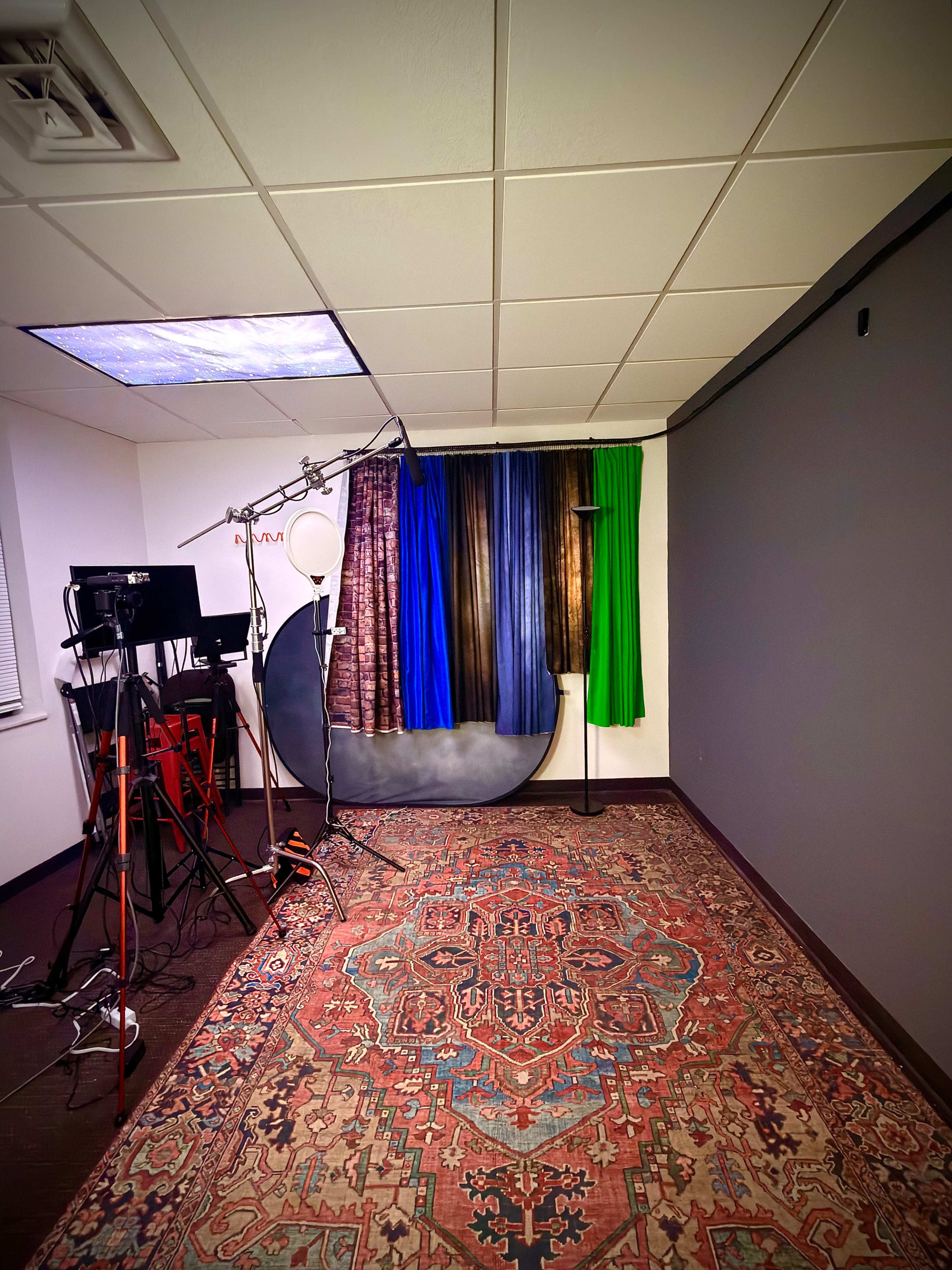 The image depicts a studio space featuring a patterned rug, a backdrop with colored curtains, and various equipment including lights and camera stands.
