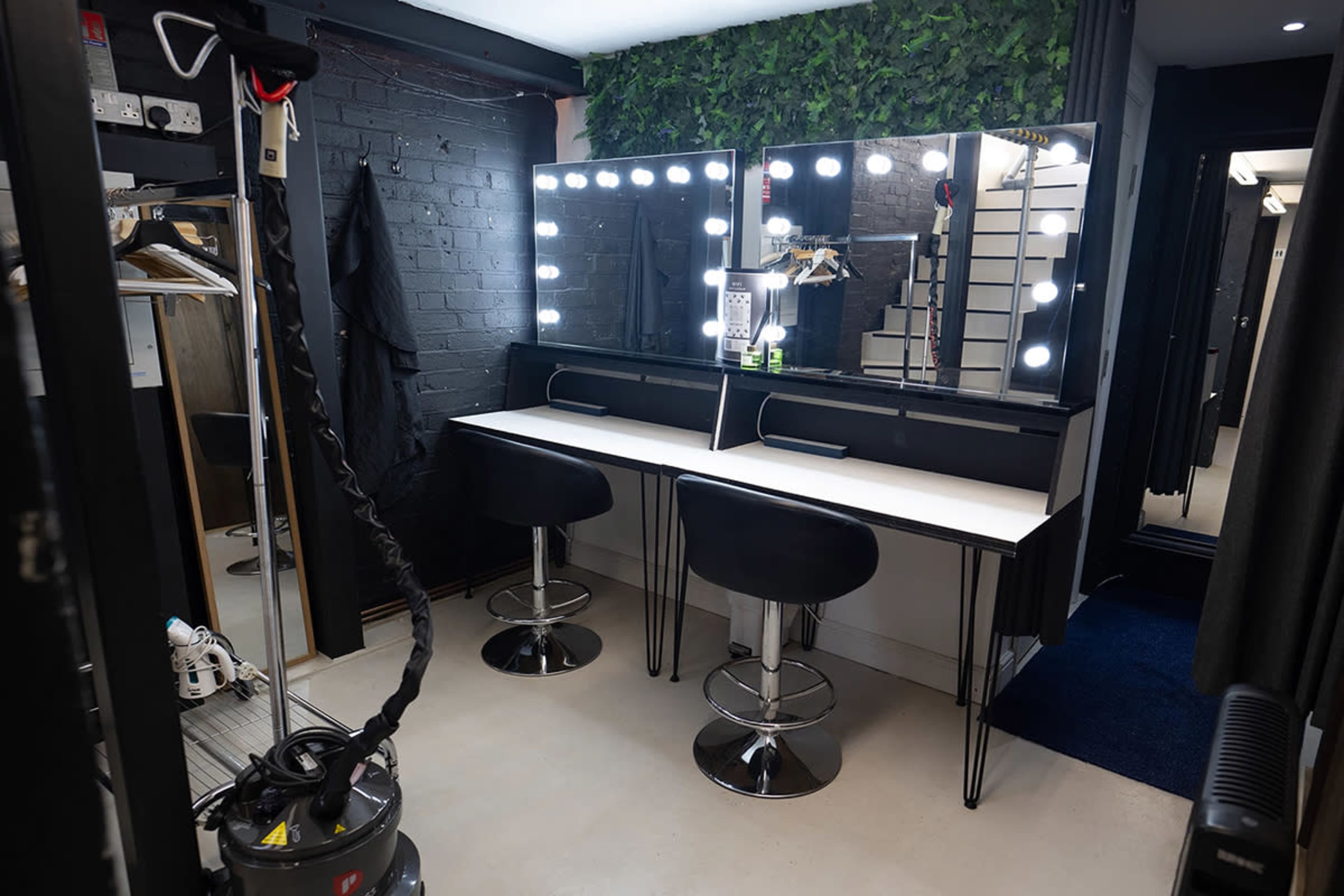 A modern styling room with two illuminated mirrors, black bar stools, a vacuum cleaner in the corner, and green foliage on the wall.