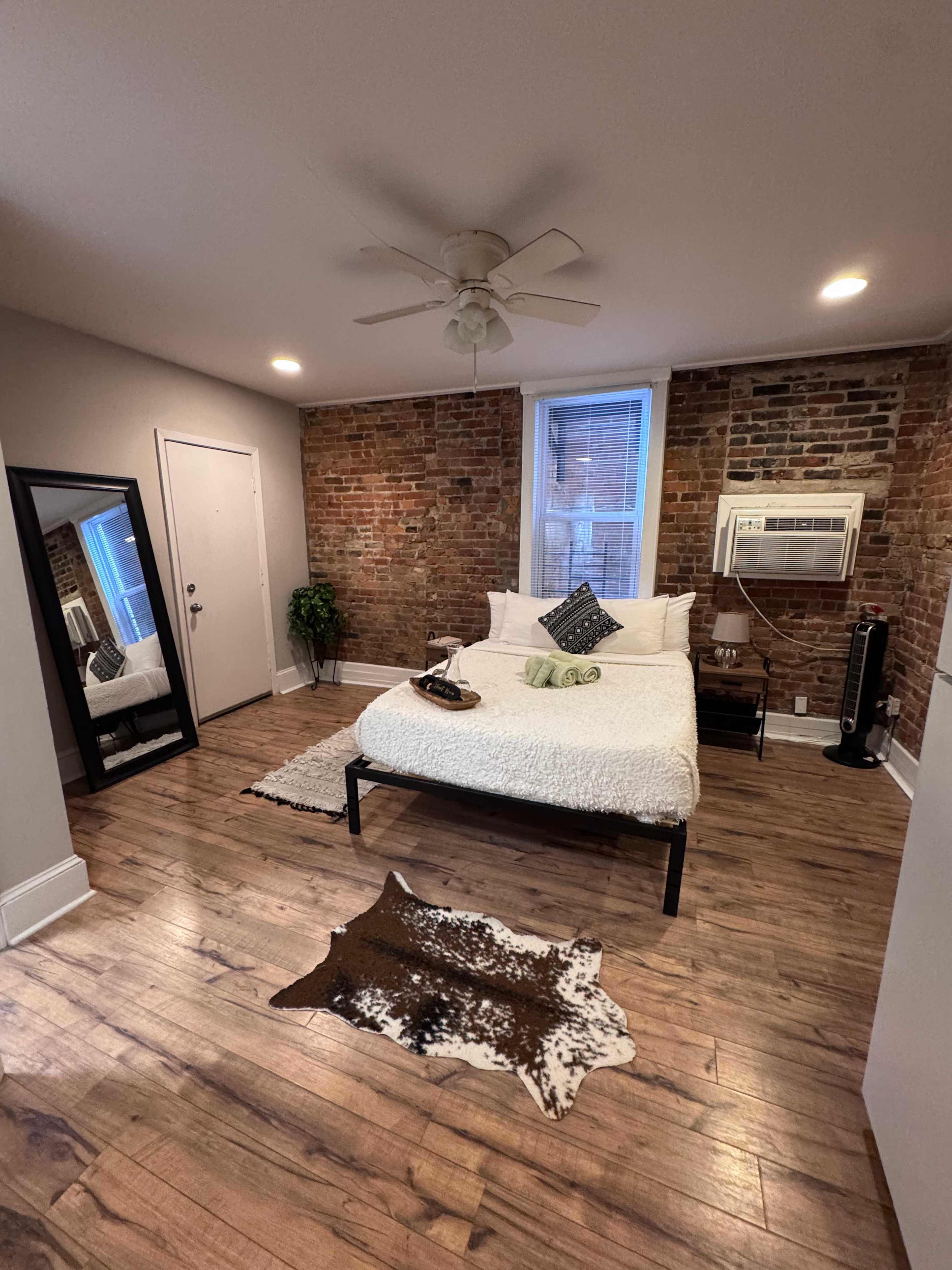 The image shows a cozy bedroom featuring a bed with white bedding, a mirror, a small plant, and a brick wall.