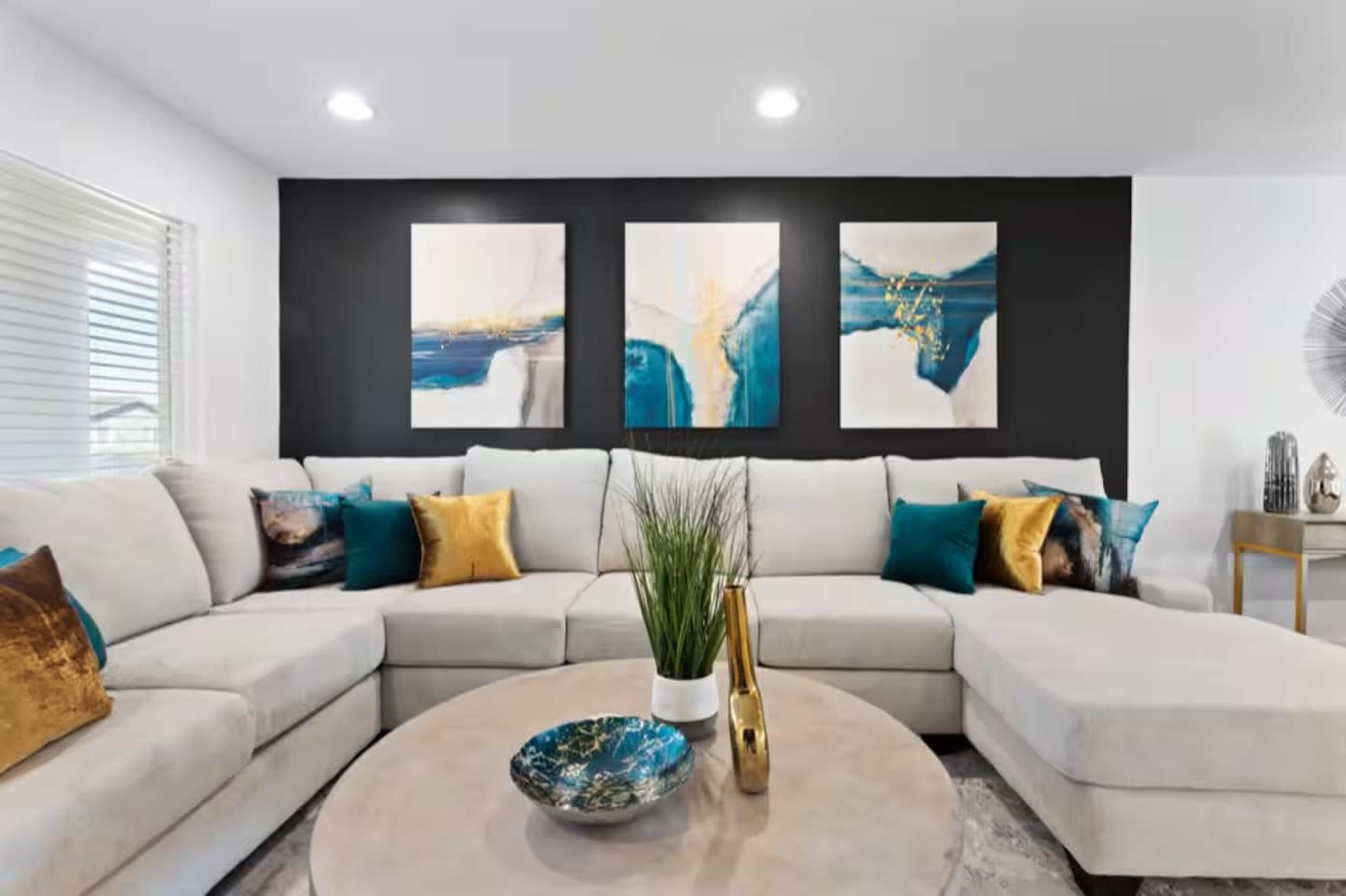 The image shows a modern living room with a light-colored sectional sofa, decorative pillows, a round coffee table, and three abstract paintings mounted on a dark accent wall.