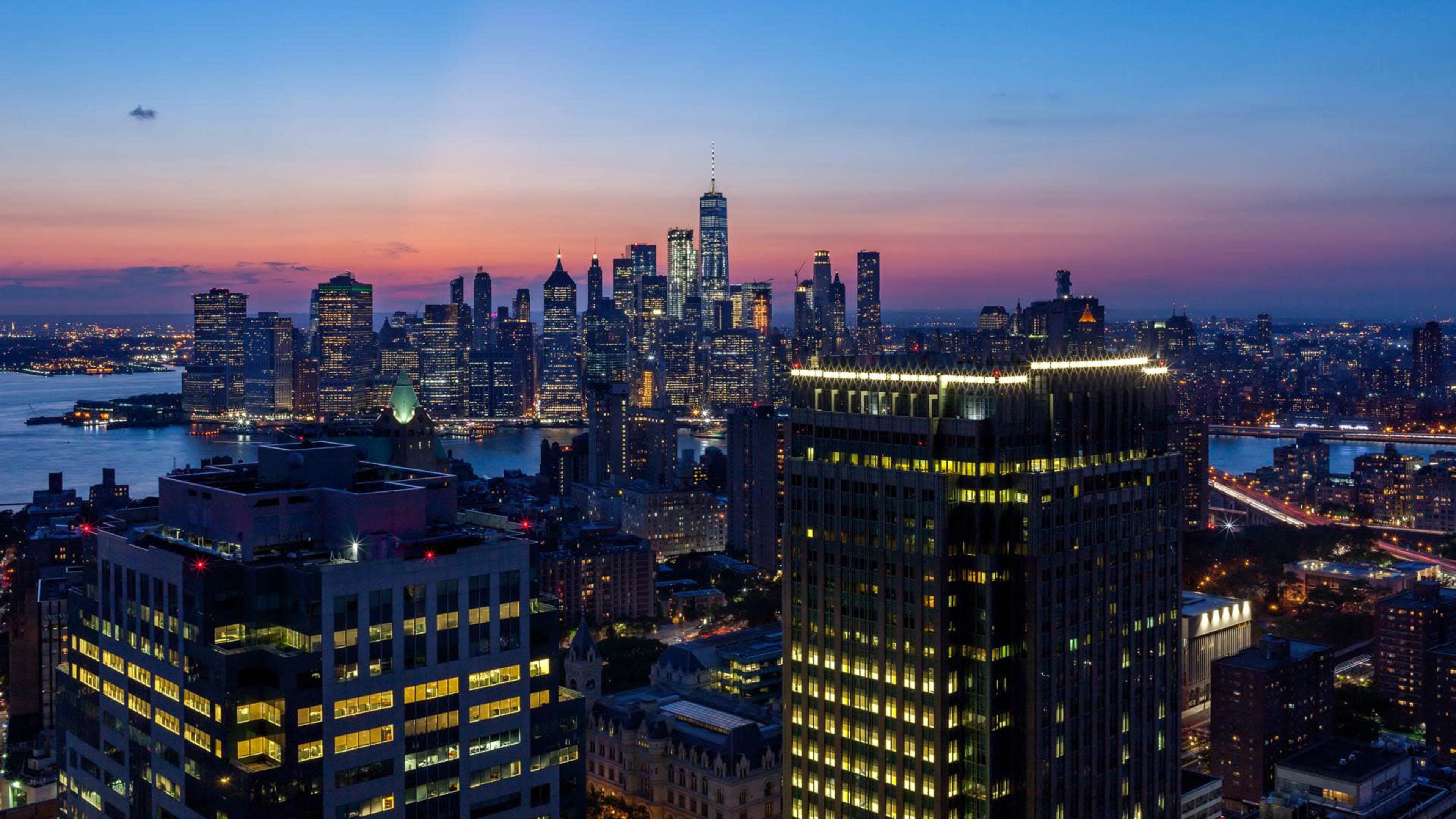 The skyline of New York City is visible at dusk, illuminated by city lights against a colorful sunset.
