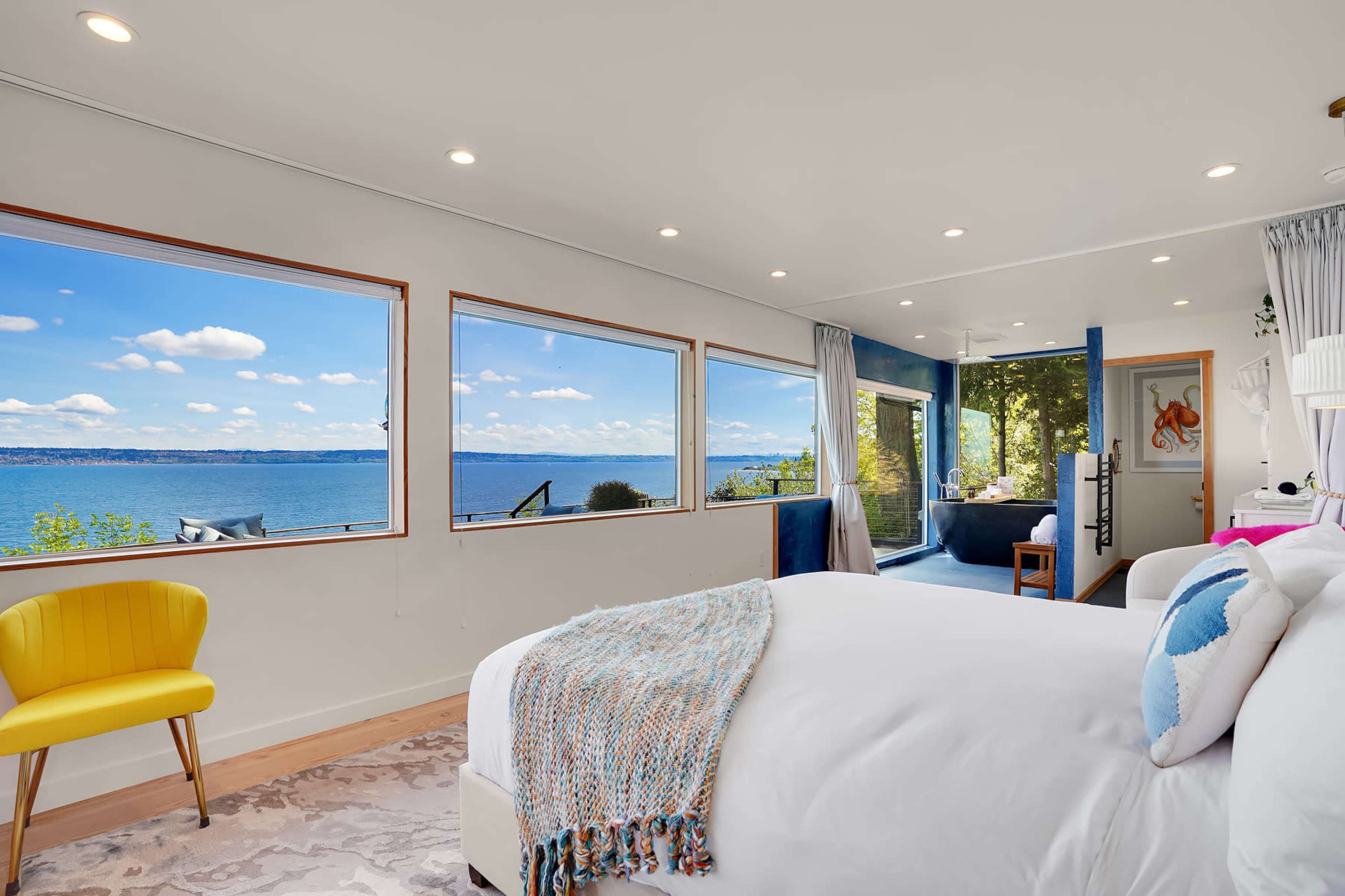 A bright bedroom features large windows with a view of the water, a bed with white linens, and a yellow chair.