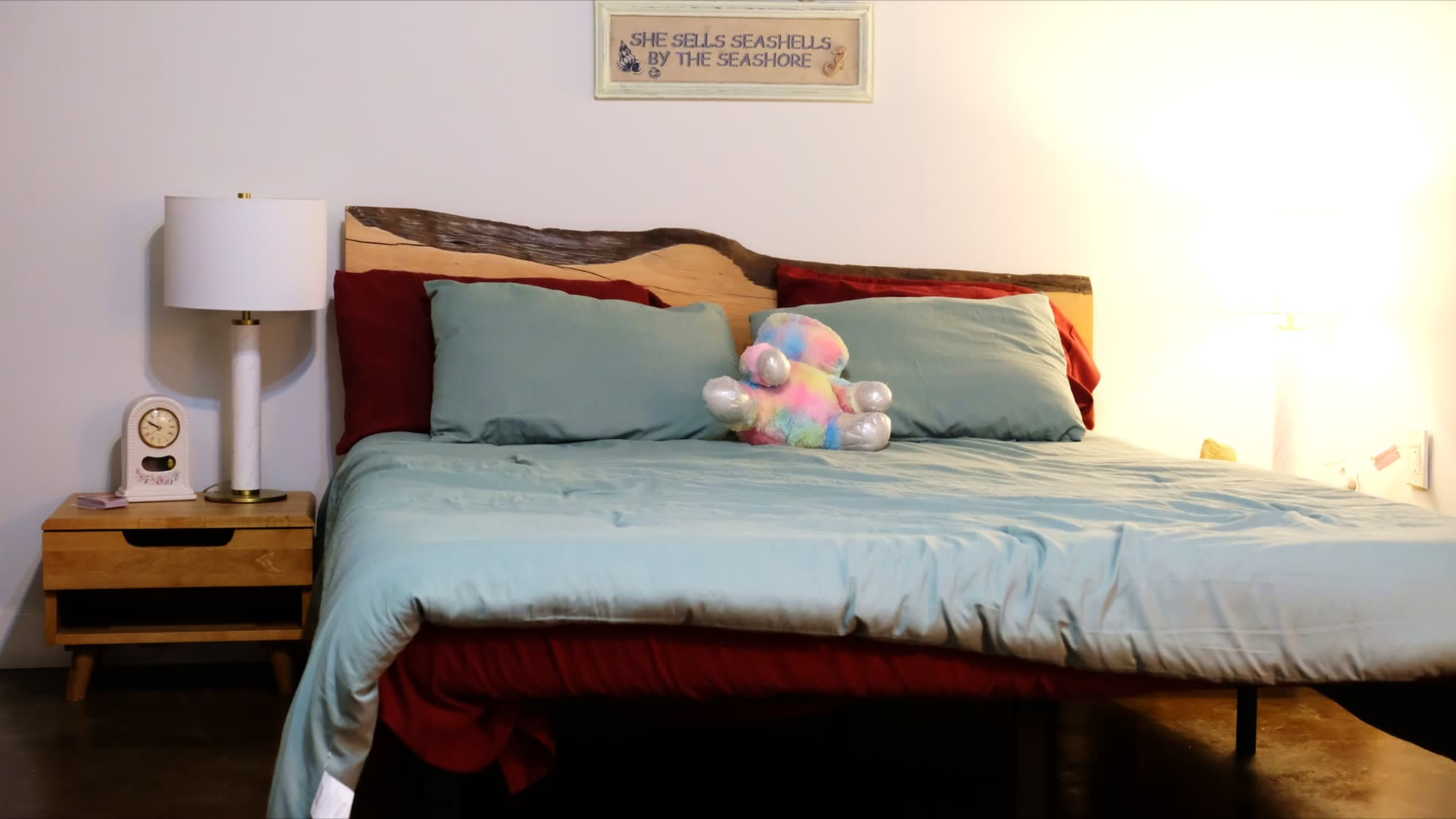 A neatly made bed with light blue and red bedding, a colorful plush toy, a bedside lamp, and a small nightstand with a clock.