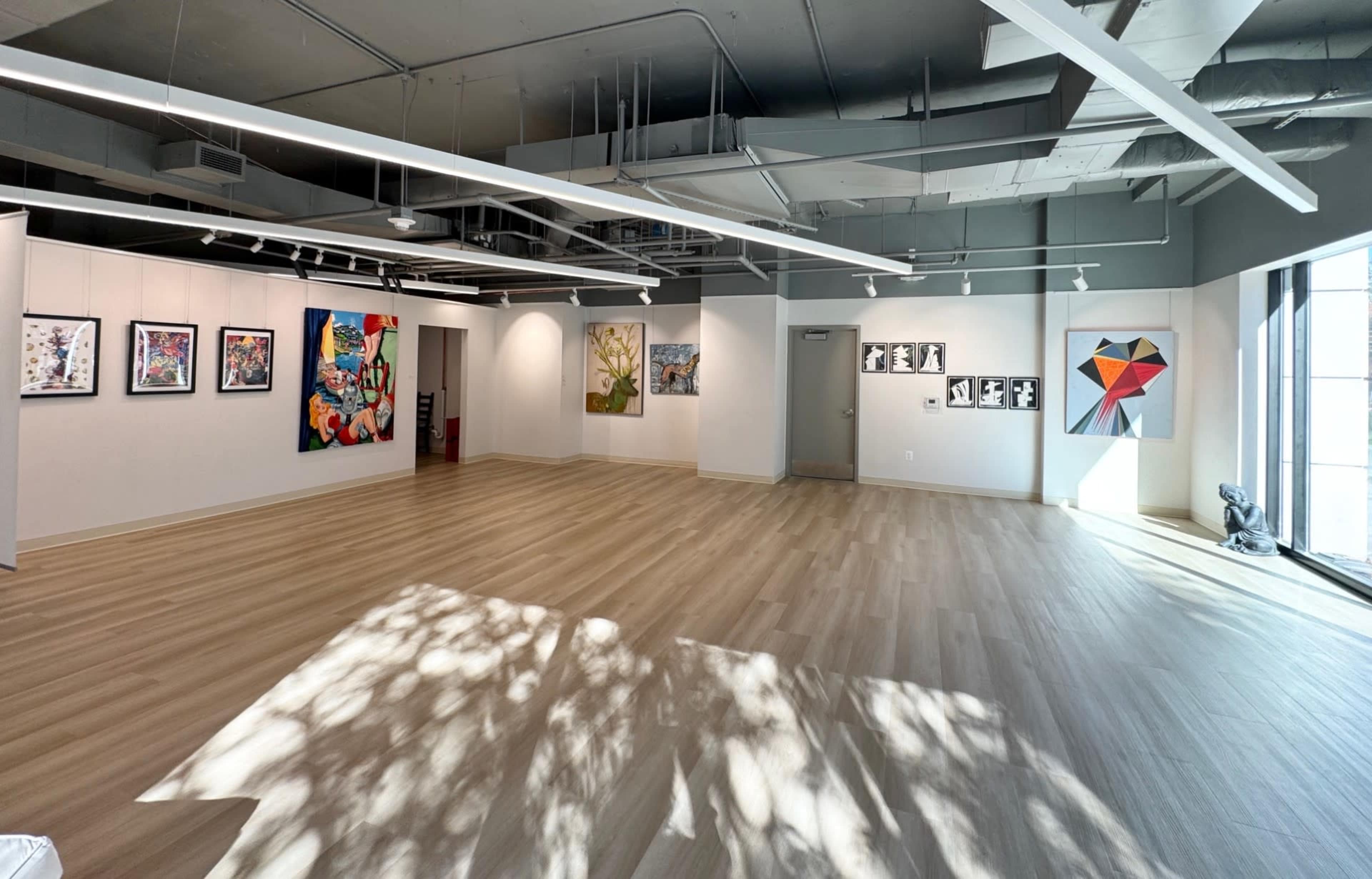 The image features an art gallery with several framed artworks displayed on white walls and a spacious wooden floor bathed in light.