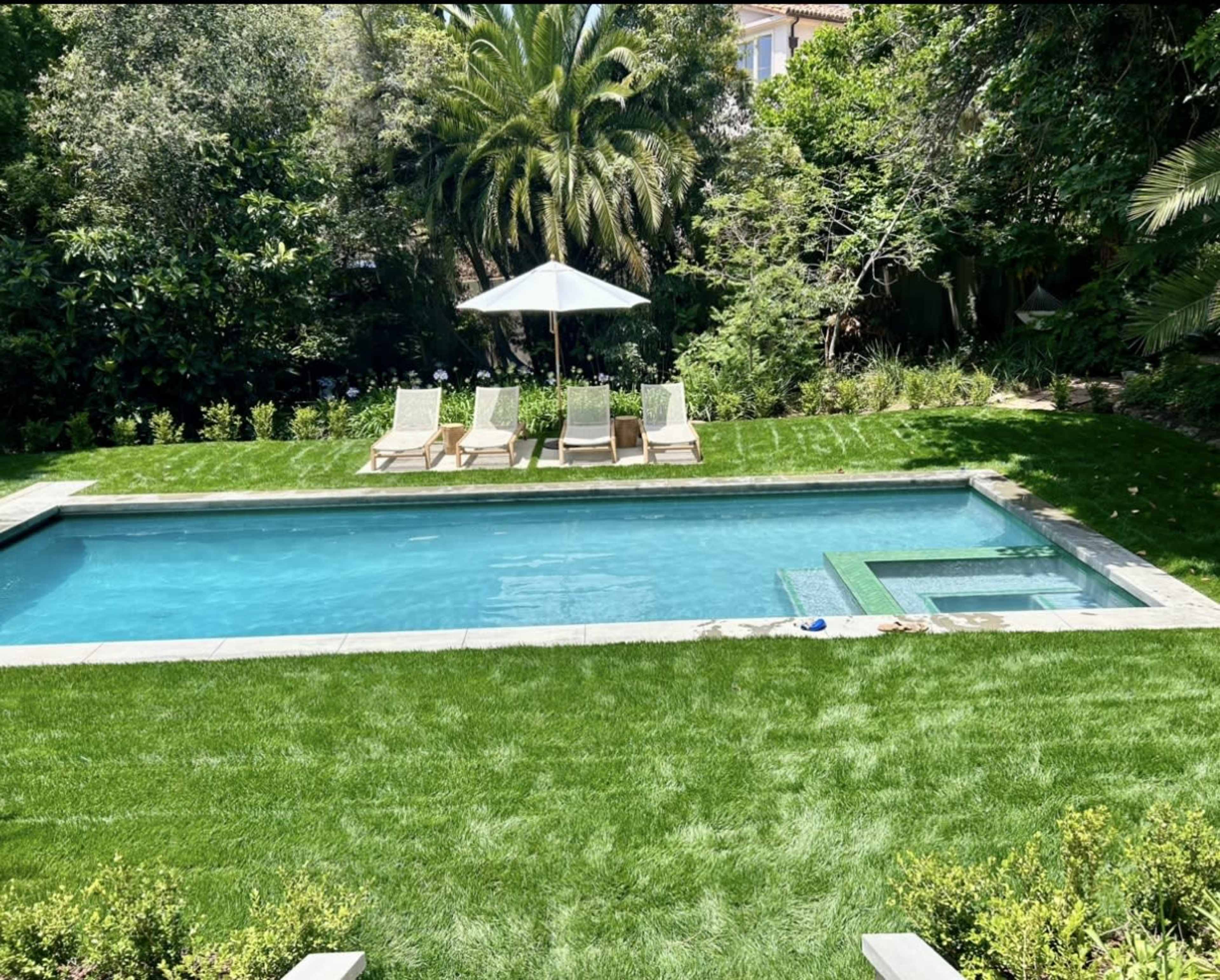 A rectangular swimming pool with four lounge chairs arranged around it, surrounded by lush greenery and a white umbrella.