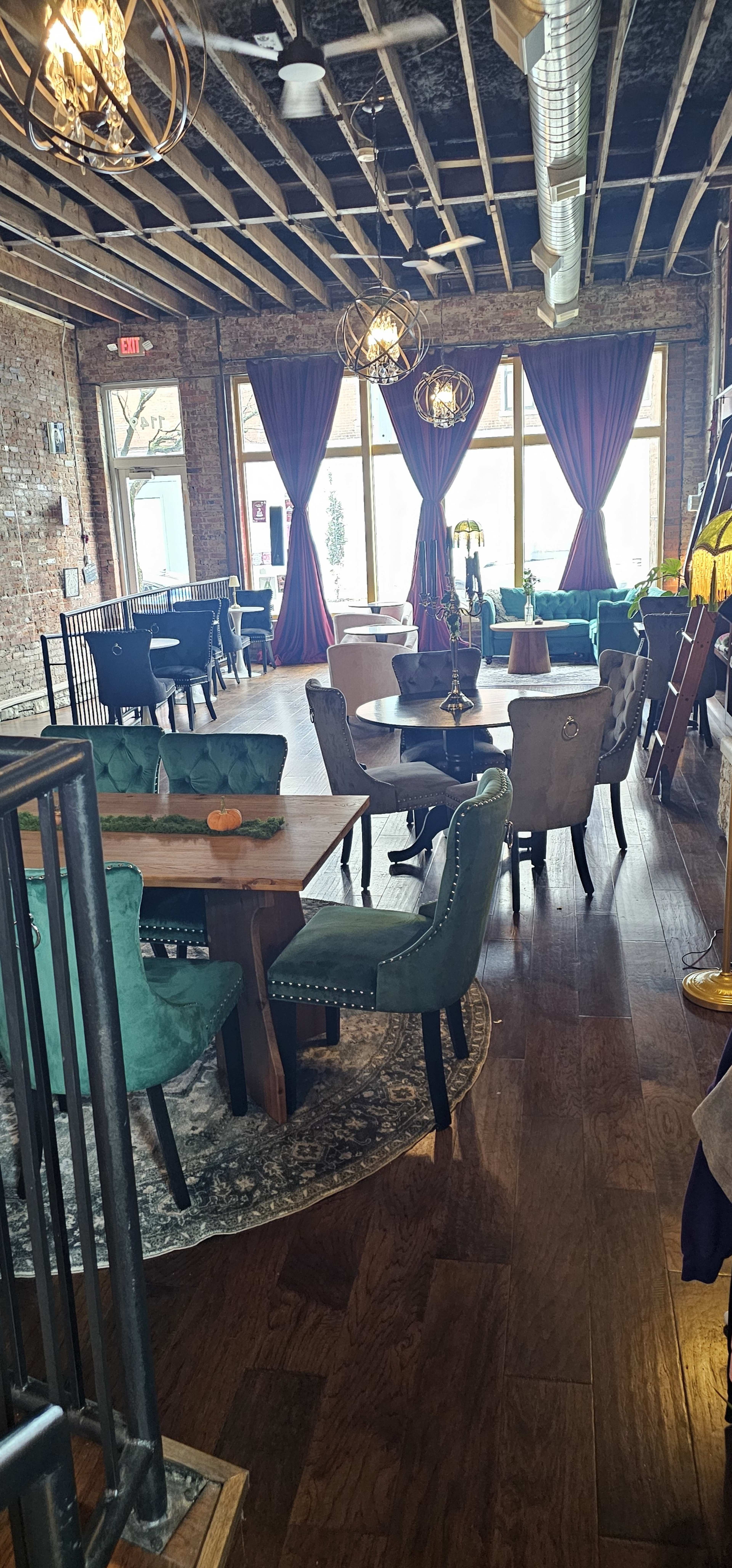The interior of a restaurant features a mix of tables and chairs with colorful upholstery, wooden floors, and large windows draped with curtains.