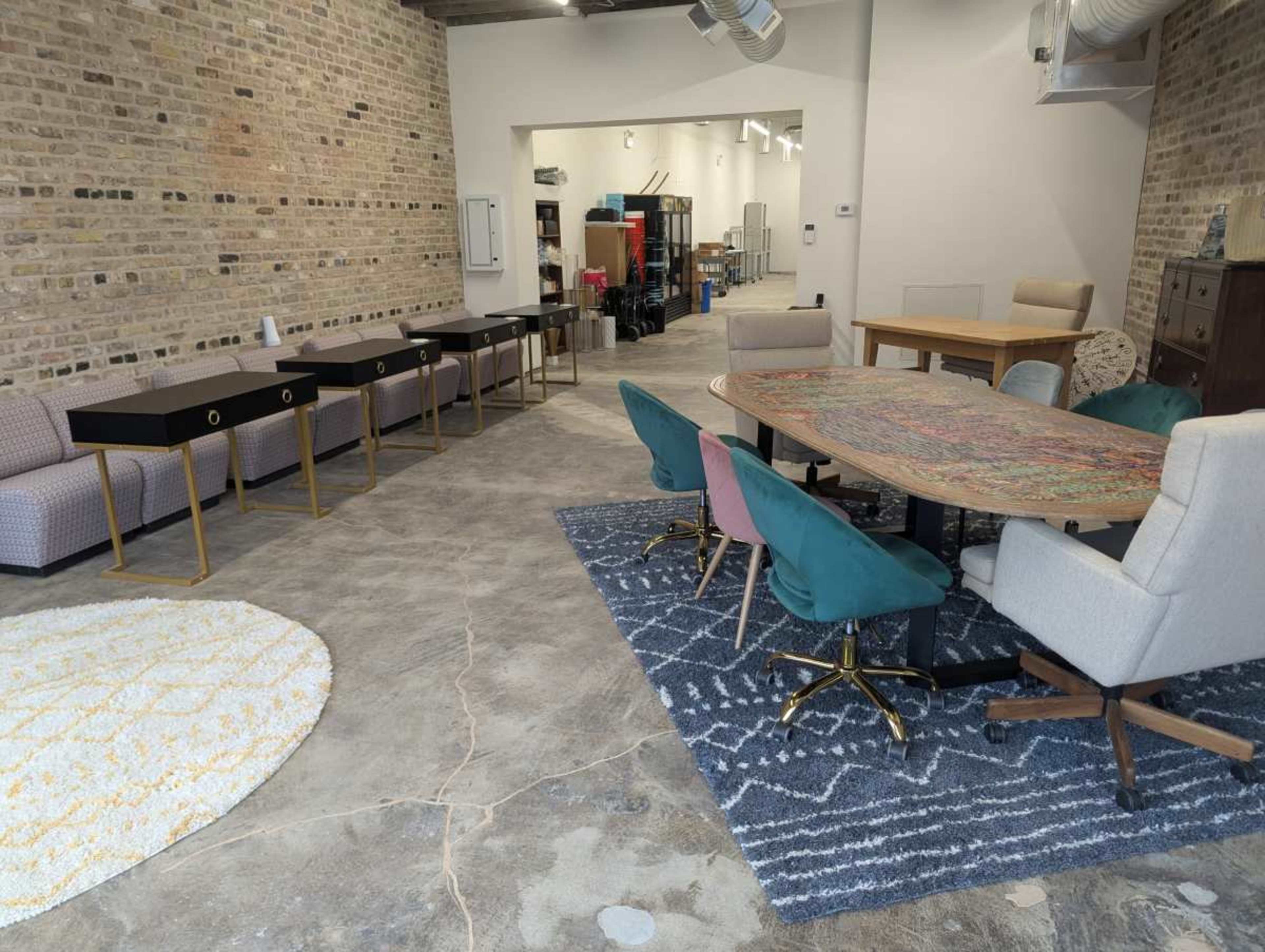 The image shows a spacious interior featuring a mix of tables and chairs, with patterned rugs on the floor and exposed brick walls.