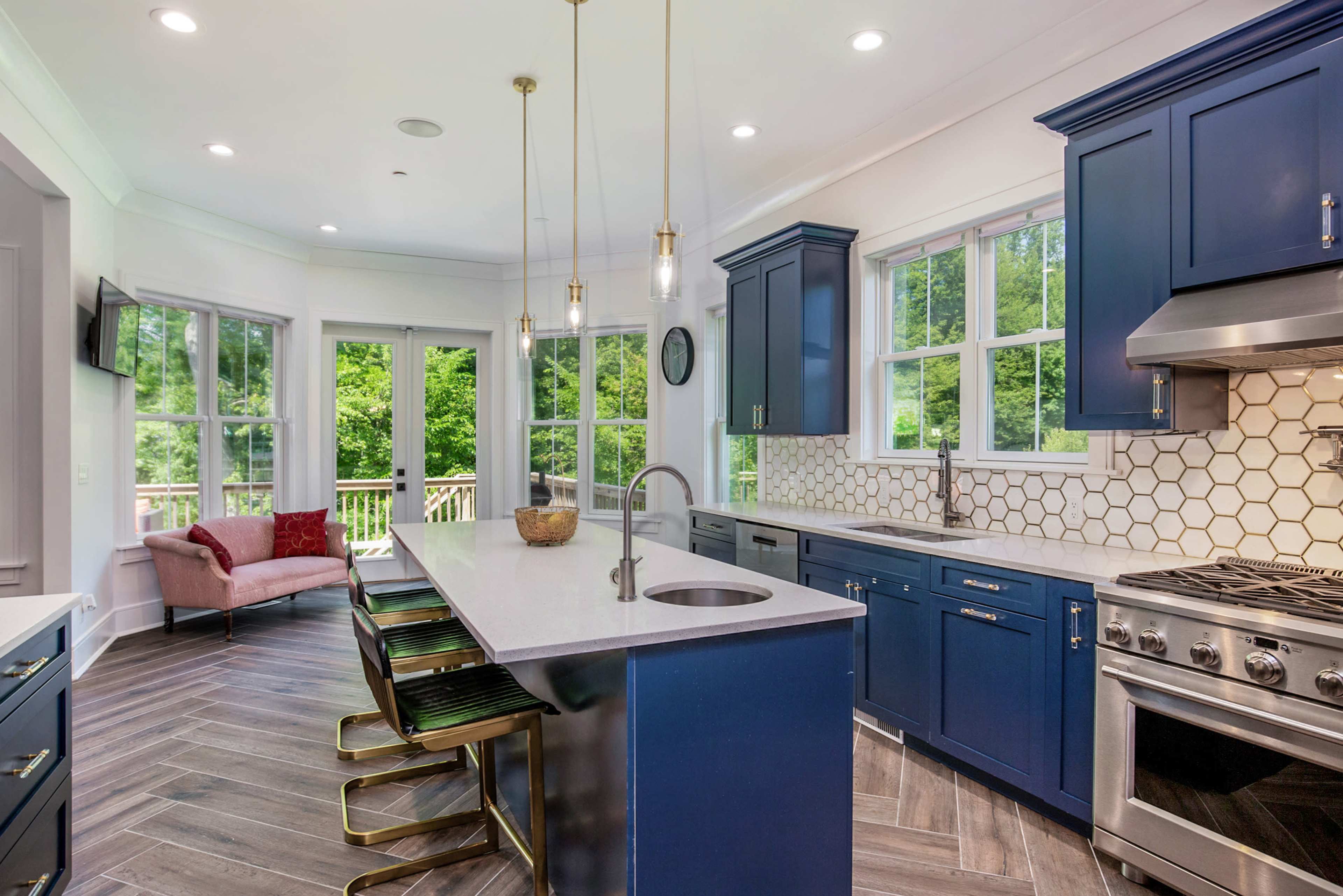 A modern kitchen features navy blue cabinets, a large island with seating, and hexagonal tile backsplash with large windows overlooking a wooded area.
