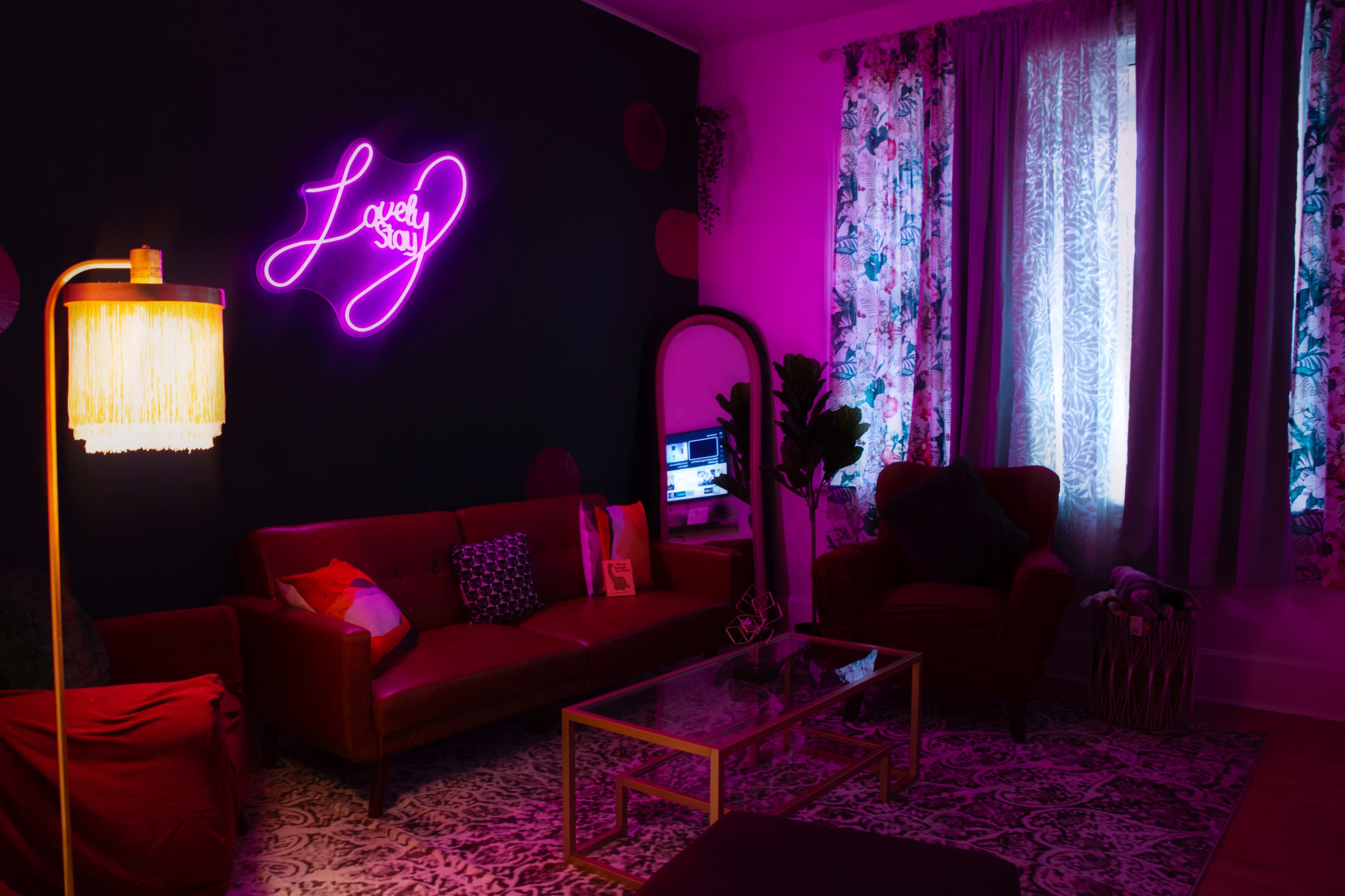 The image features a stylish living room with pink neon lighting, a red velvet sofa, and large windows adorned with patterned curtains.