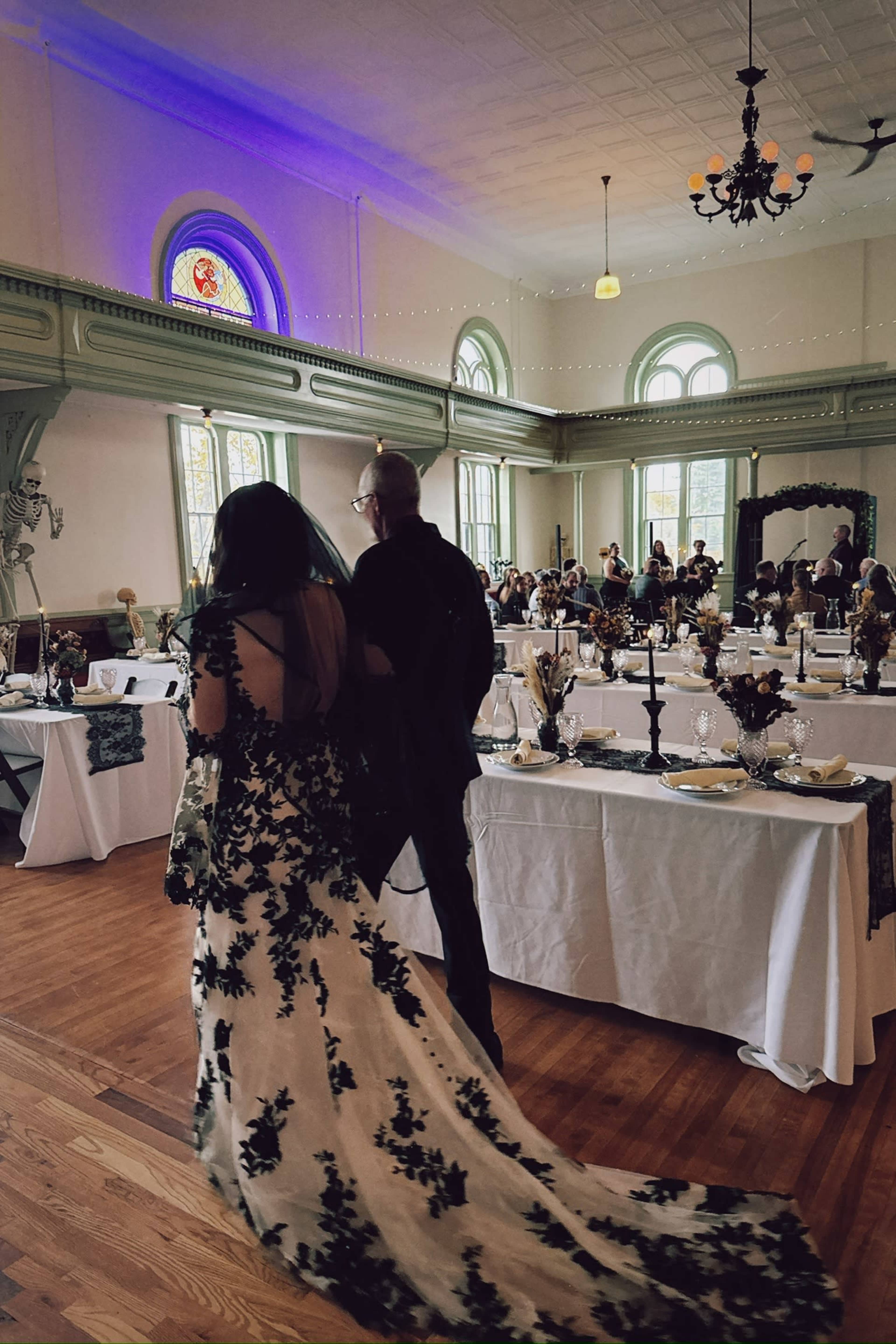 A woman in a floral-patterned gown walks down the aisle toward a decorated reception area filled with guests and tables set for a celebration.