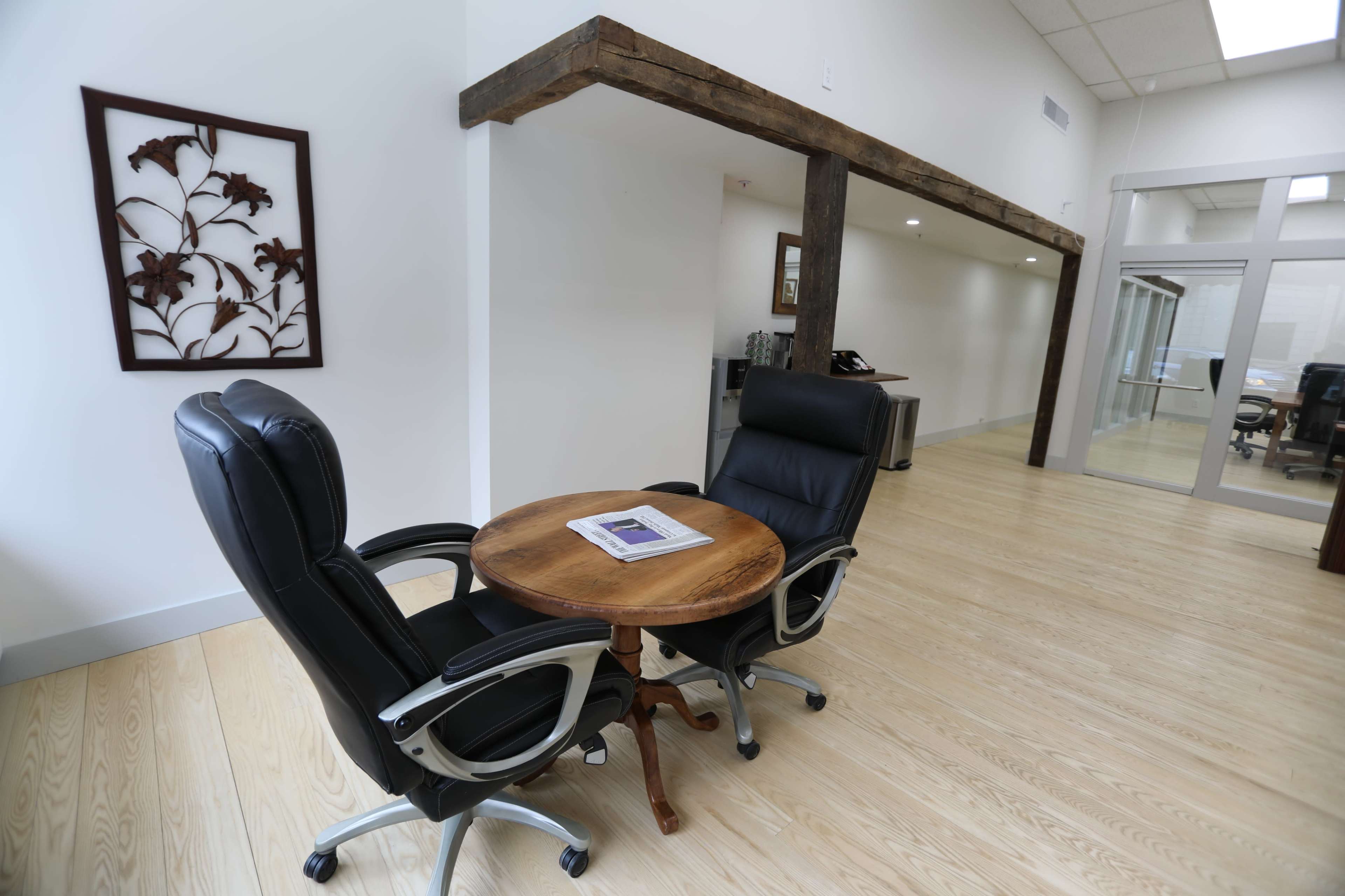 A modern office space featuring two black ergonomic chairs positioned around a circular wooden table on a light wooden floor.