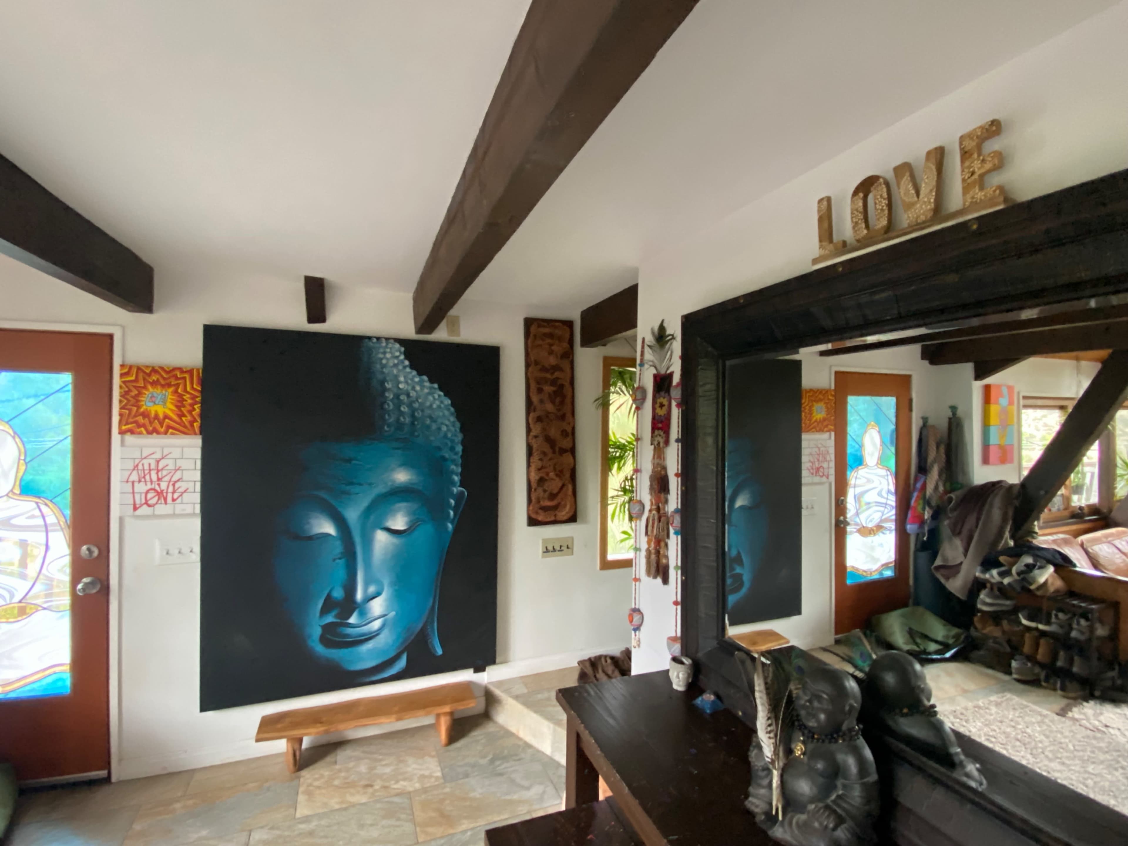 A large mural of a serene Buddha face hangs on the wall of a room with wooden beams, colorful stained glass doors, and various decorative items.