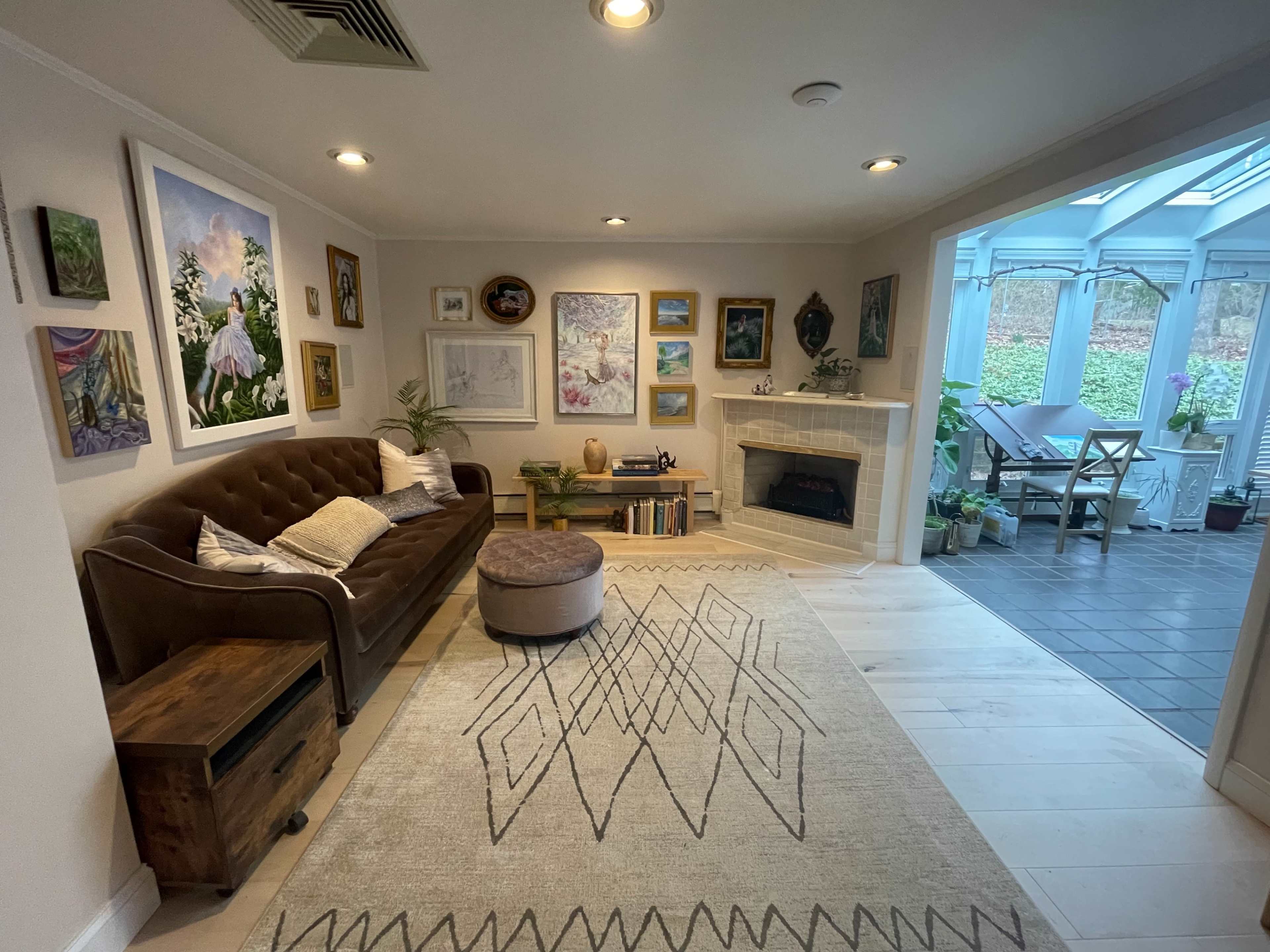 A cozy living room featuring a brown couch, a round ottoman, a large area rug with a geometric pattern, and various artworks on the walls, adjoining a sunlit conservatory.