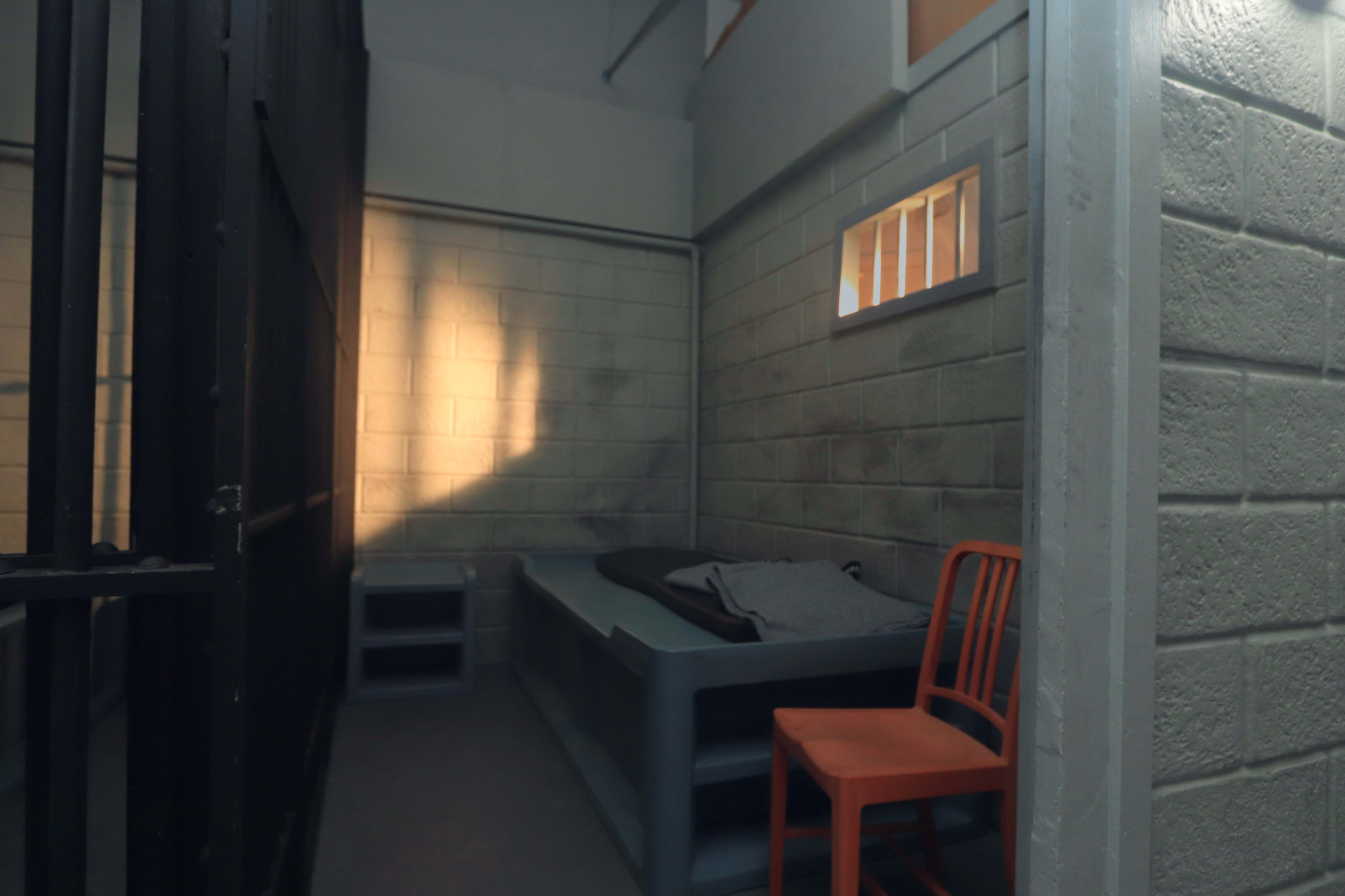The image shows the interior of a small, sparse jail cell with a bed, a chair, and a barred entrance.