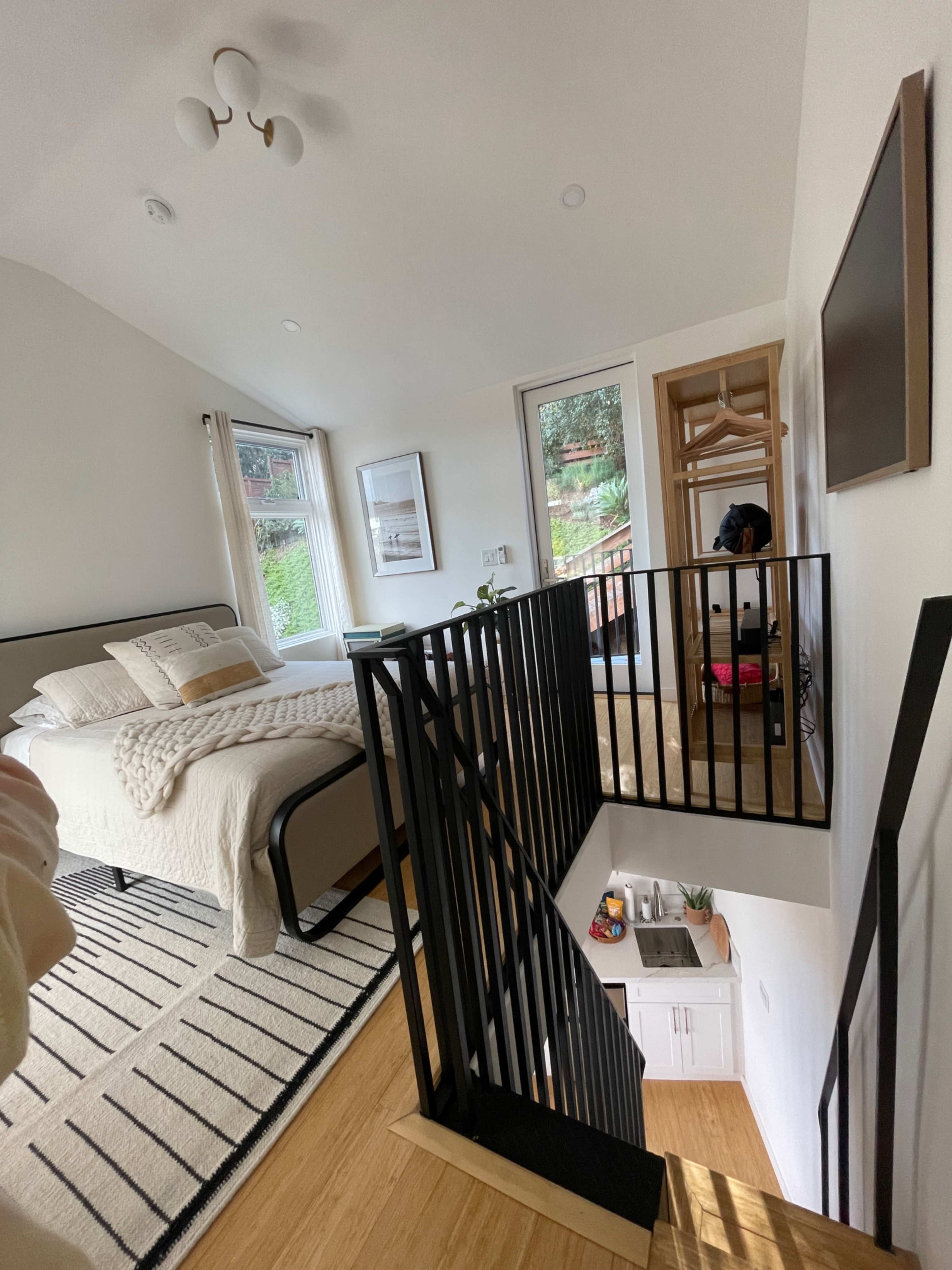 The image shows a well-lit bedroom with a bed, a staircase with a black railing, and a window overlooking a garden.