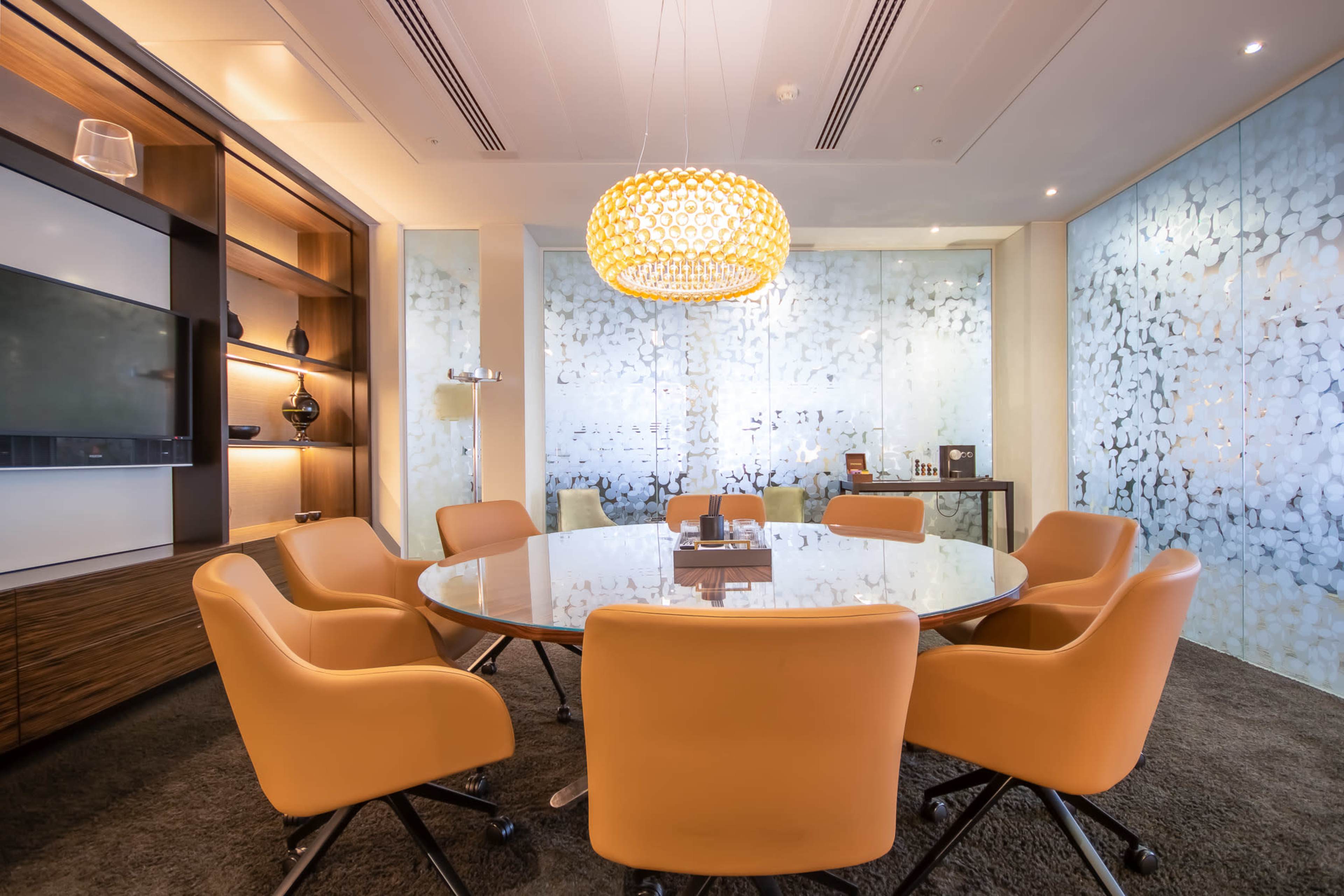 A modern conference room features a round glass table surrounded by orange chairs, with a decorative pendant light and frosted glass walls.