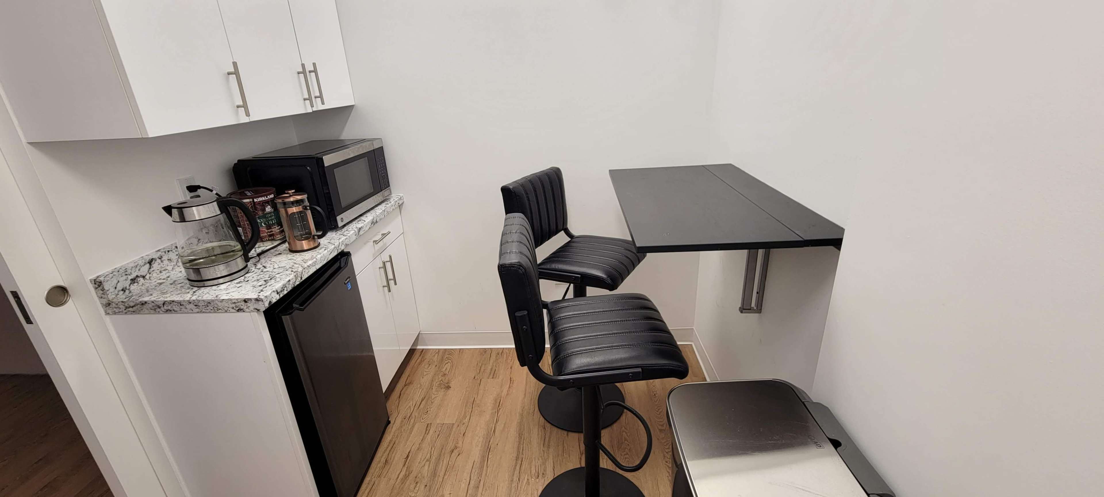 A small kitchenette featuring a black countertop with two stools, a microwave, a mini fridge, and a metal trash can.