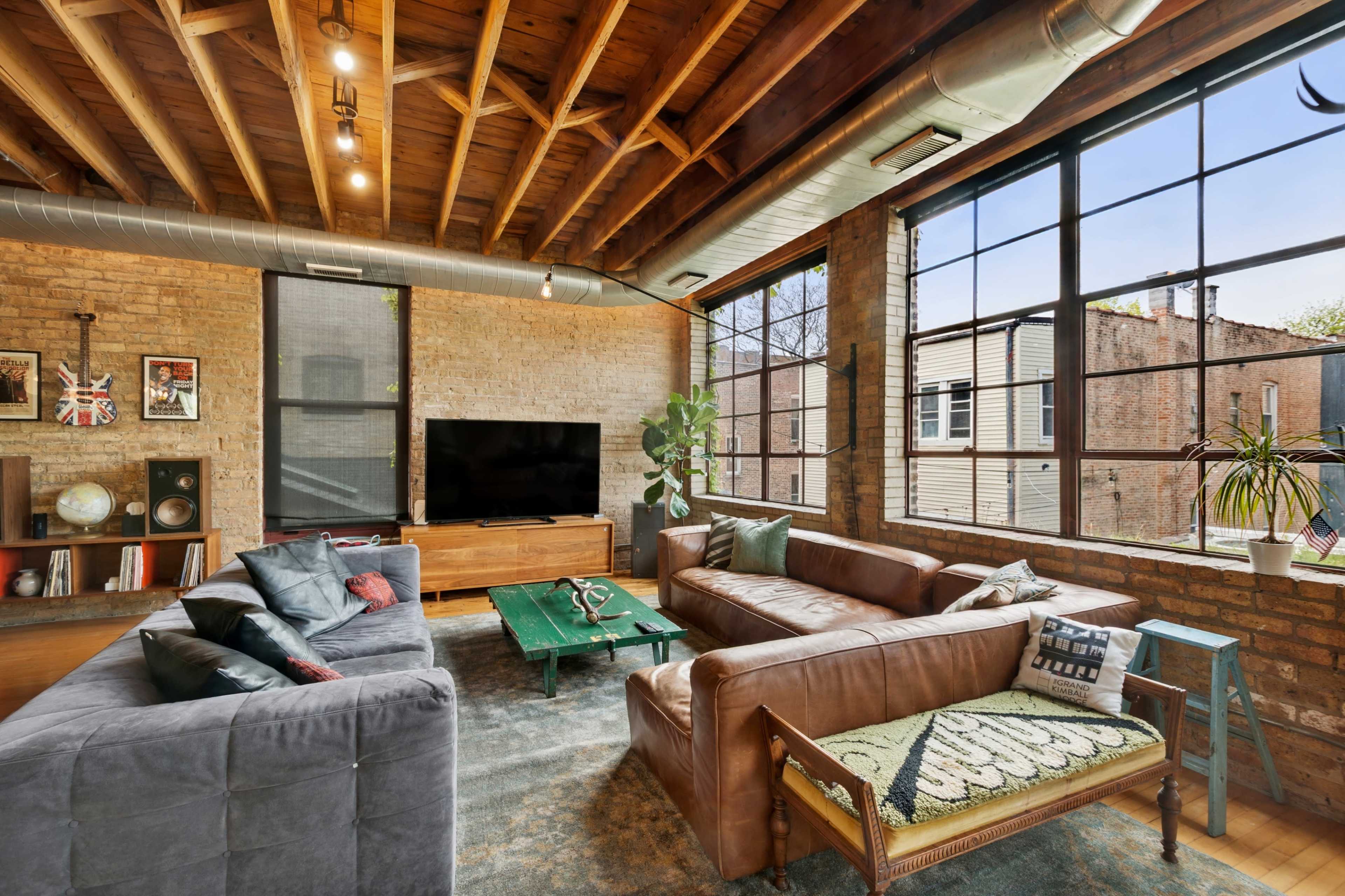 The image shows a spacious living room with exposed wooden beams, large windows, and a combination of modern and rustic furniture.