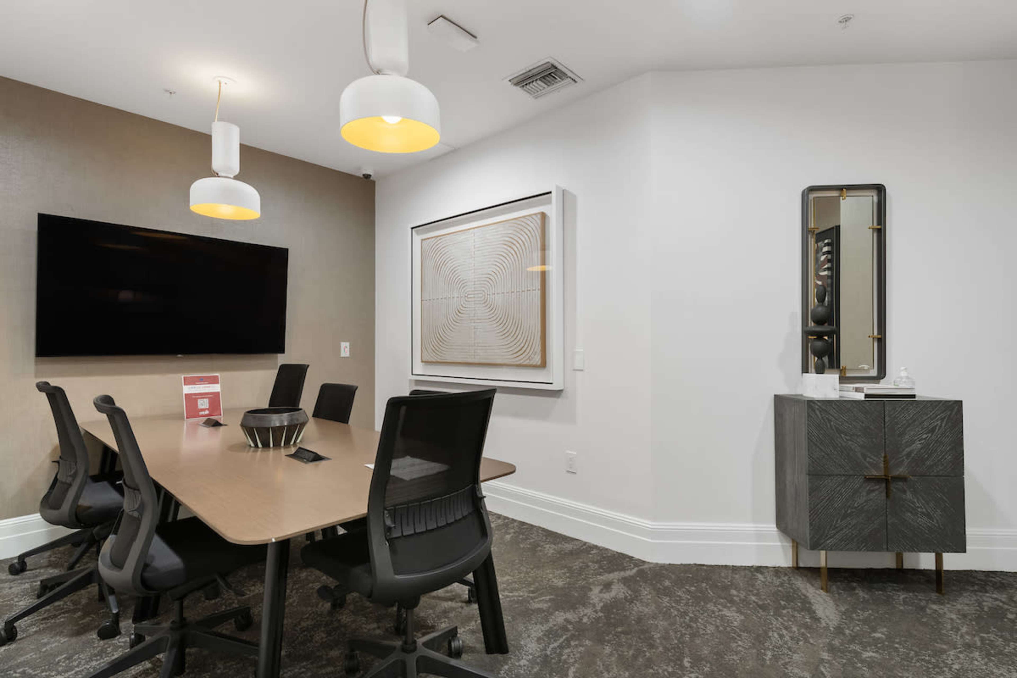 A modern conference room features a large table surrounded by ergonomic chairs, a wall-mounted television, and decorative art on the adjacent wall.