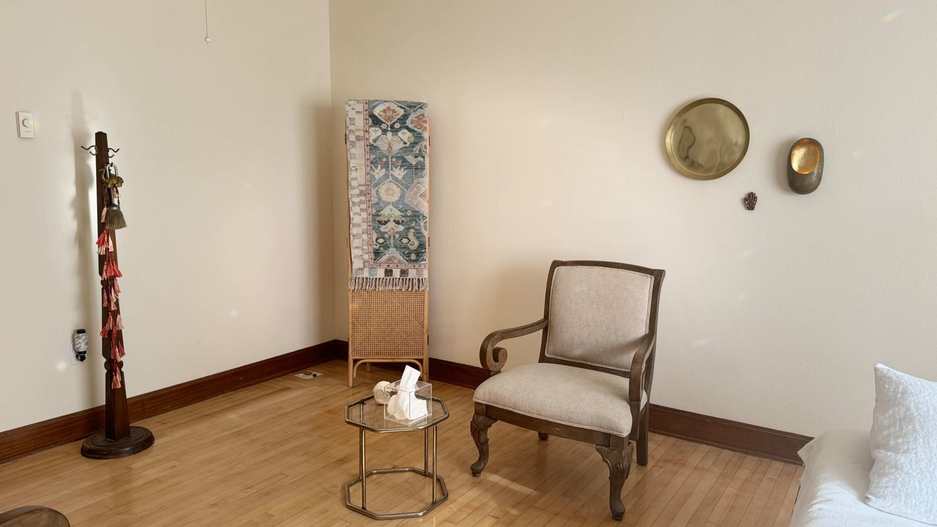 The image shows a minimalist room featuring a wooden chair, a small glass table with tissue, a decorative piece against the wall, and a wall-mounted mirror.