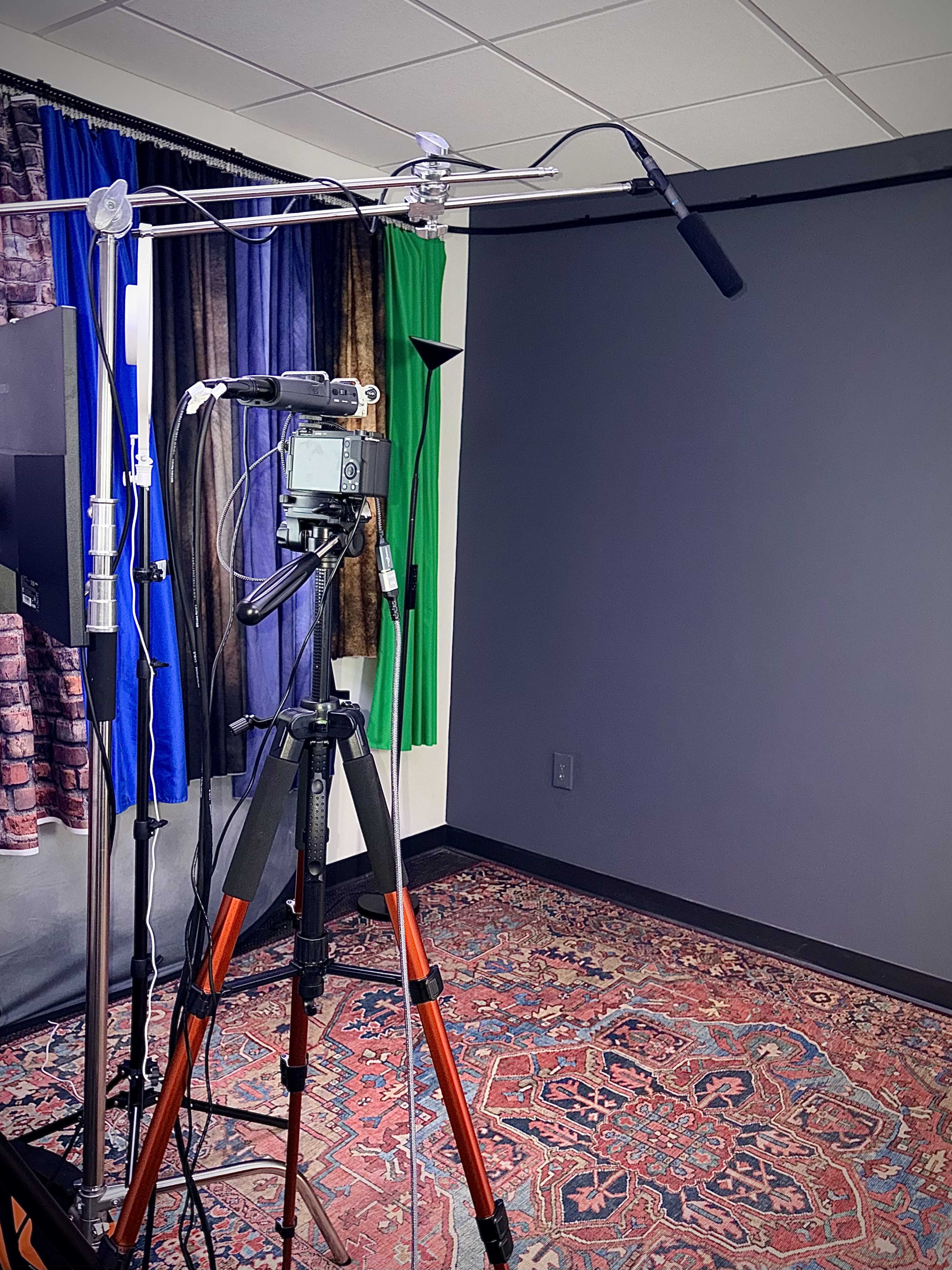 A camera mounted on a tripod is set up in a room with a plain gray wall and colorful fabric backdrops.