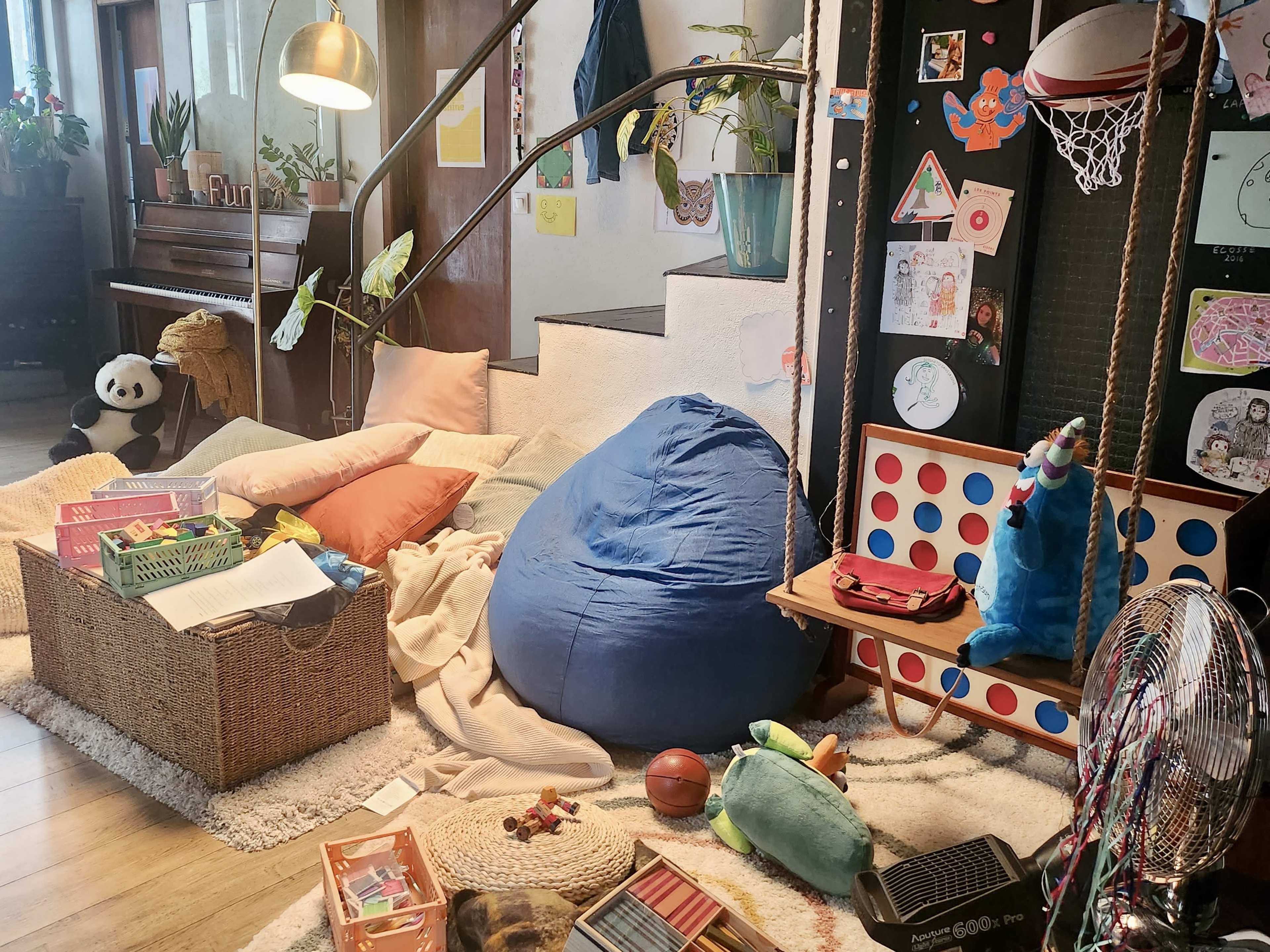 A cozy, cluttered play area featuring a blue bean bag, scattered toys, and a staircase in a sunlit room with plants and a piano in the background.