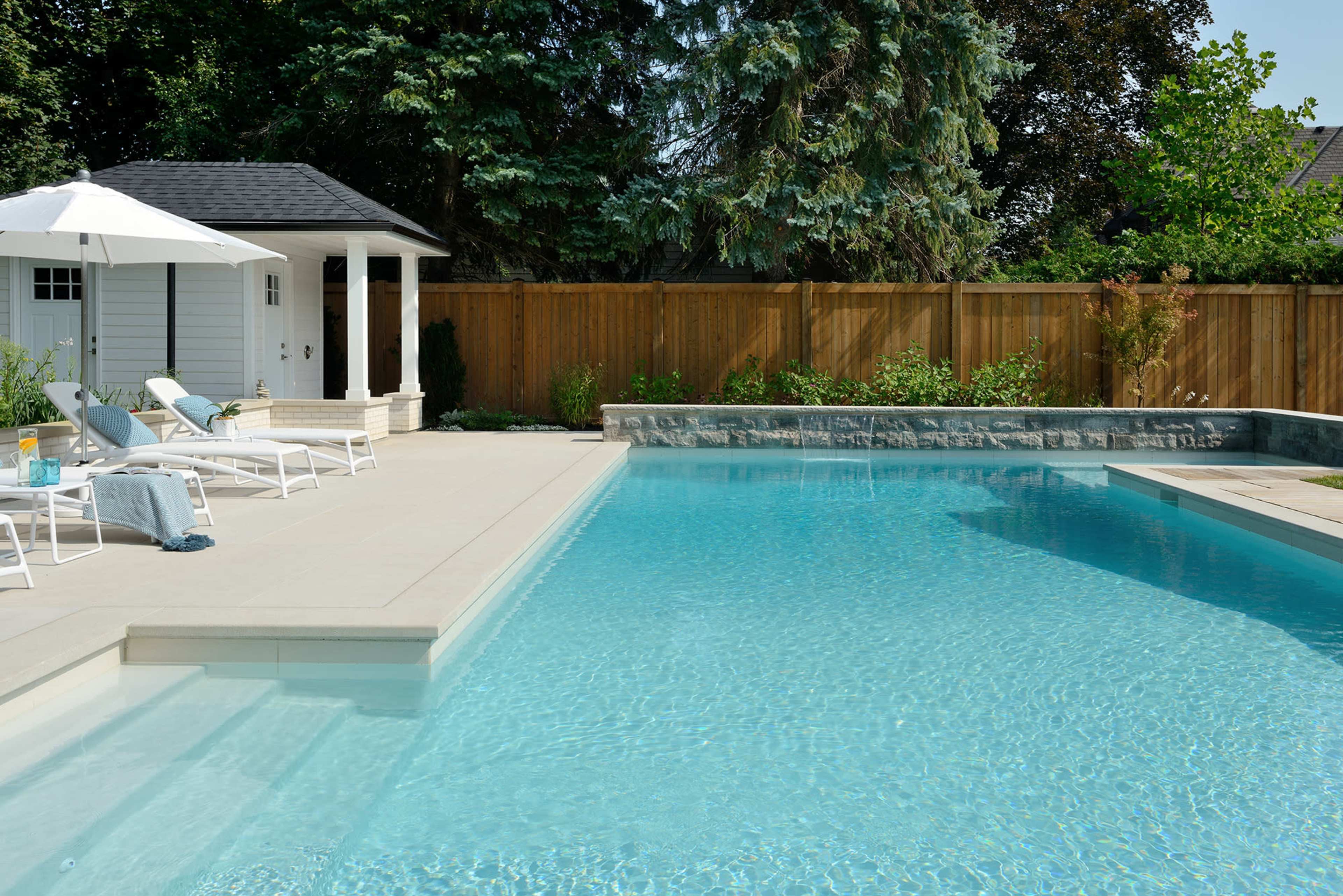 A clear swimming pool surrounded by lounge chairs and an umbrella, with a gazebo and wooden fence in the background.