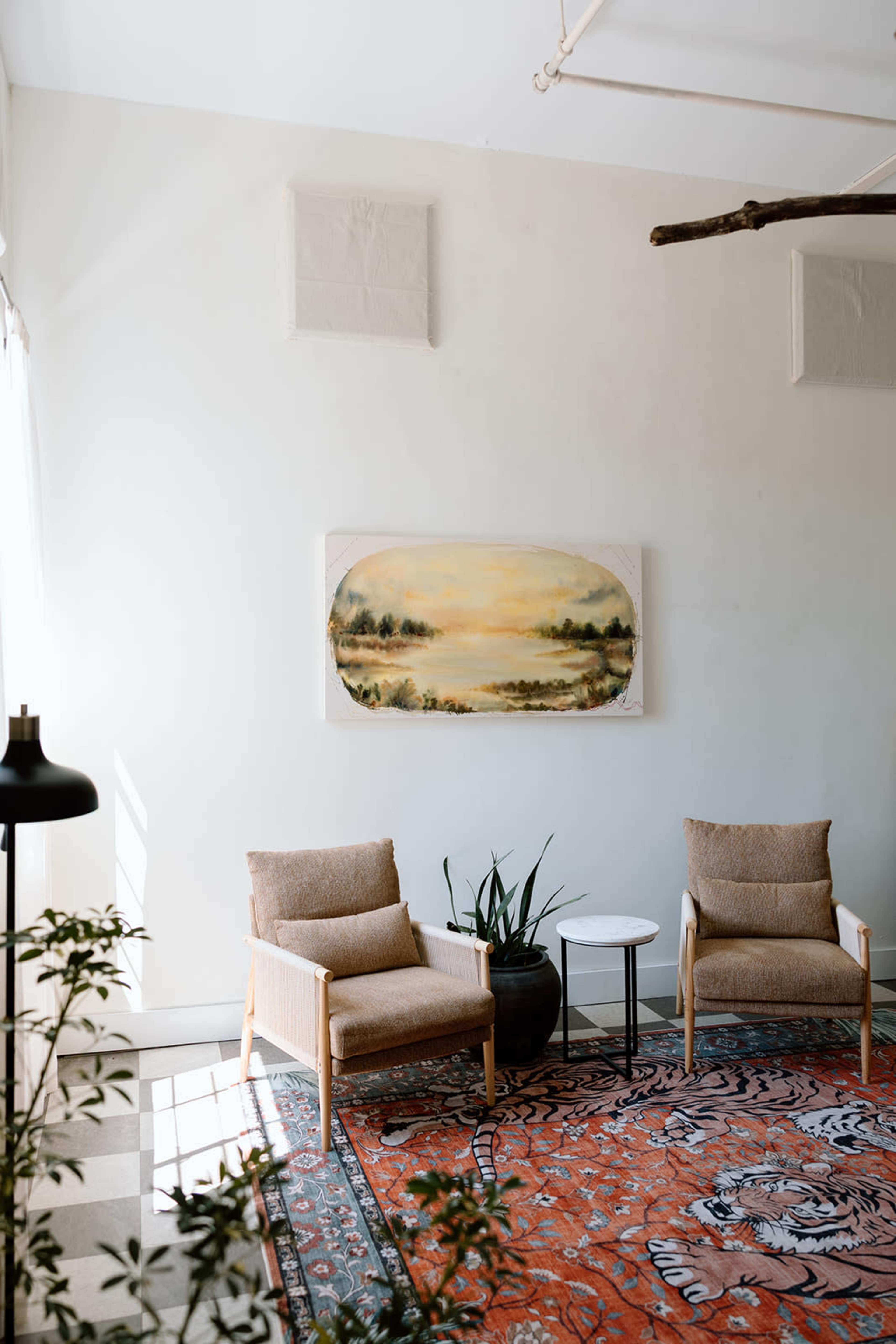 The image shows a sunlit corner of a room with two beige armchairs, a small round table, a potted plant, and a large landscape painting on the wall above a patterned rug.