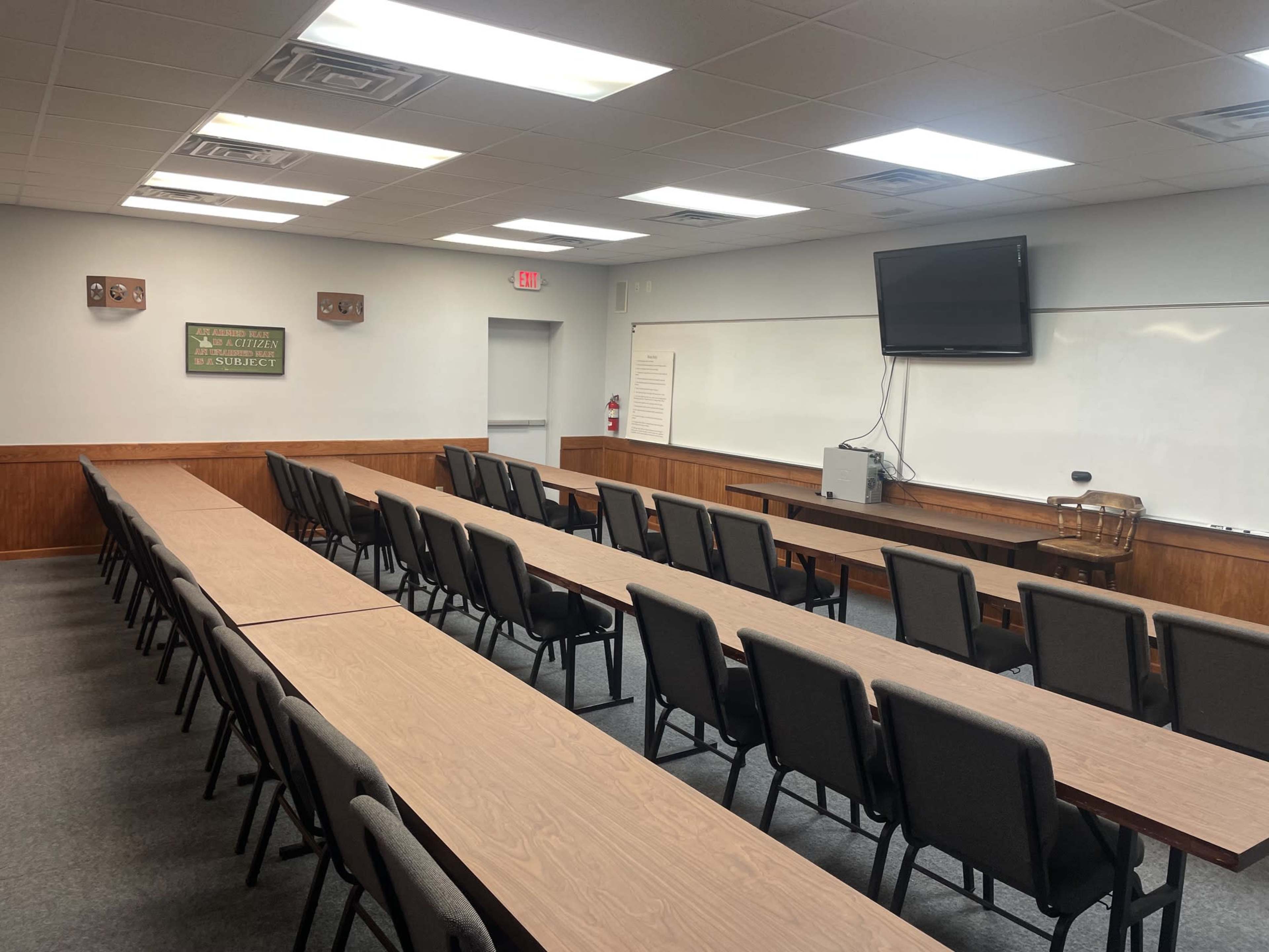 The image shows a classroom or meeting room with rows of tables and chairs, a whiteboard, and a television screen mounted on the wall.