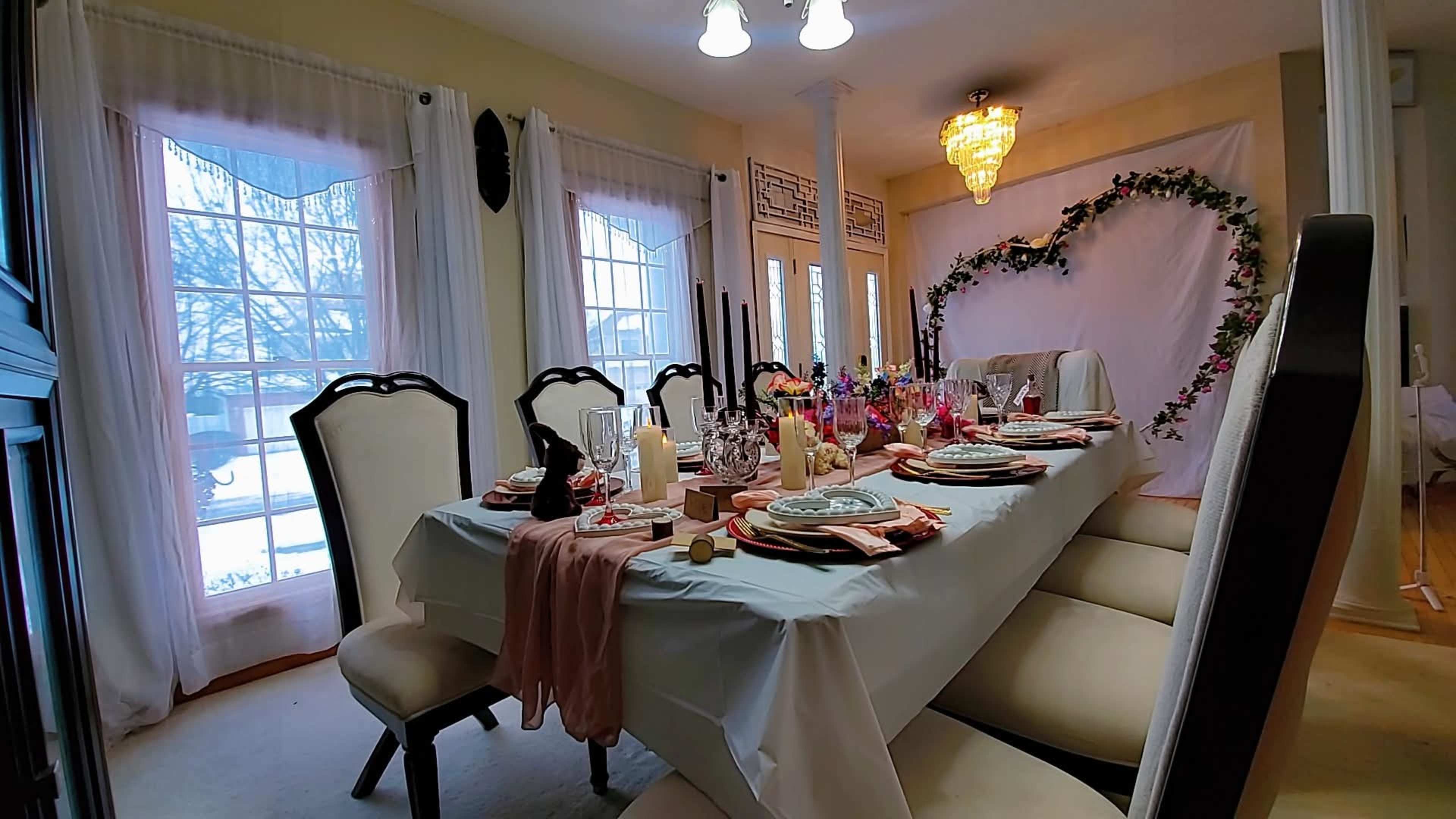 A dining table is elegantly set for a meal, adorned with decorative plates, utensils, candles, and floral arrangements, while large windows allow natural light into the room.