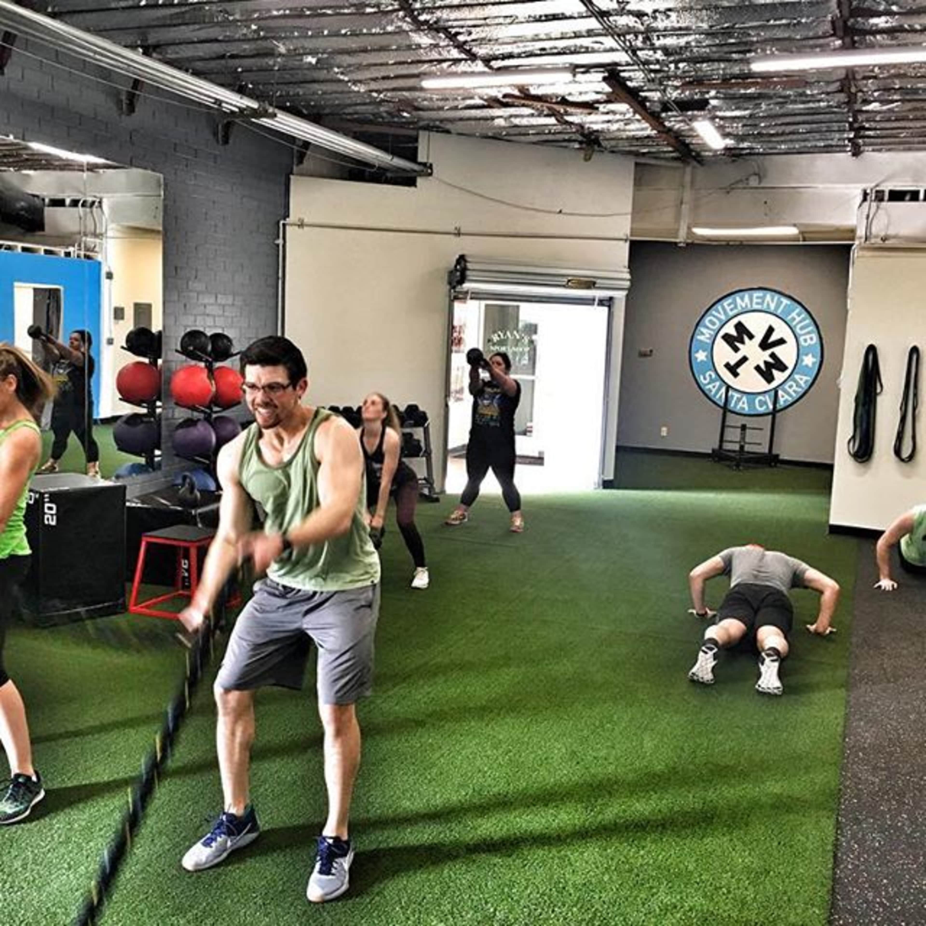 A group of individuals is engaged in various fitness activities in a gym, with a focus on strength training and conditioning exercises.