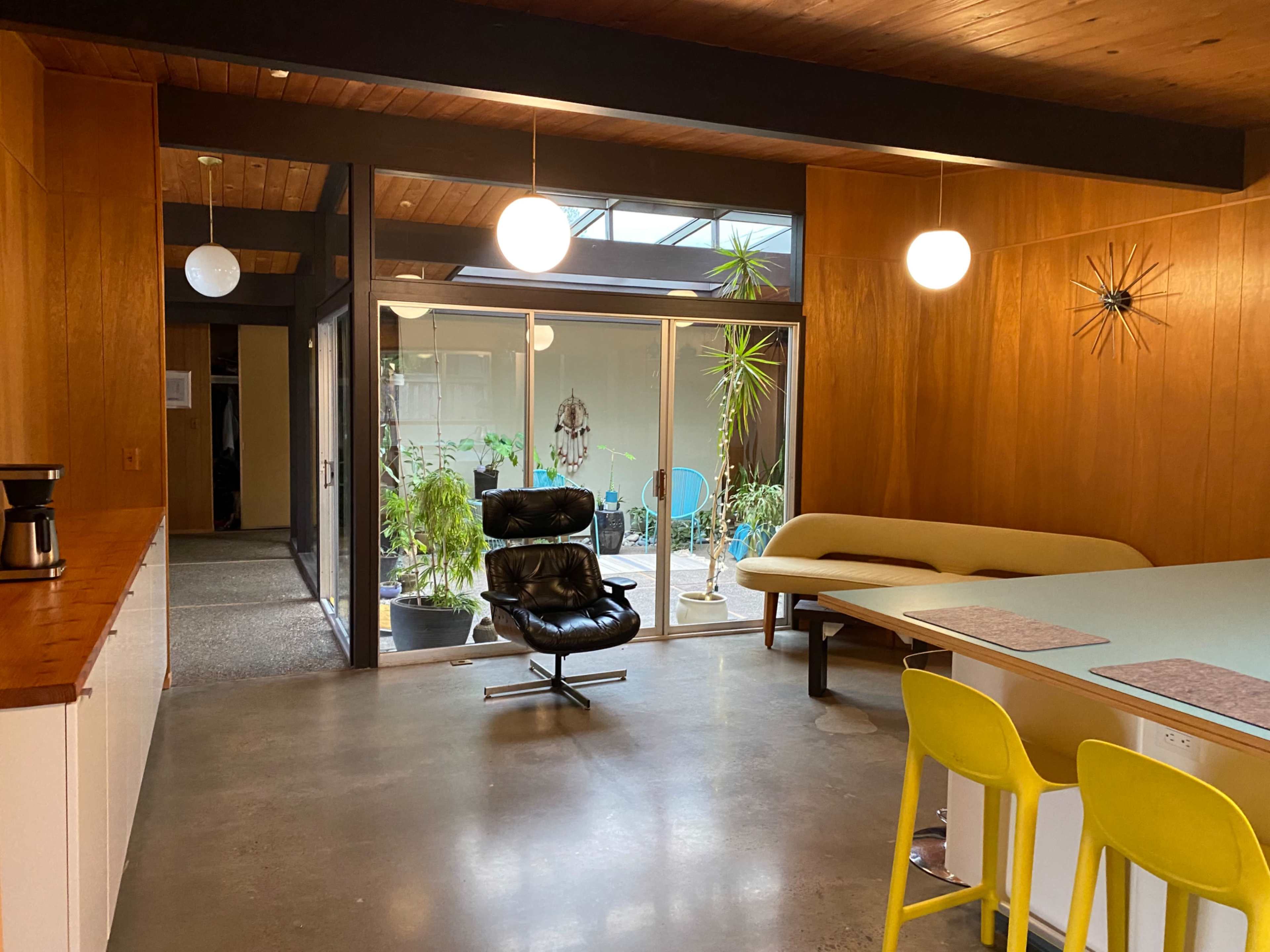 A modern interior space with a black lounge chair, a light-colored sofa, and a dining table, all surrounded by indoor plants and large windows.