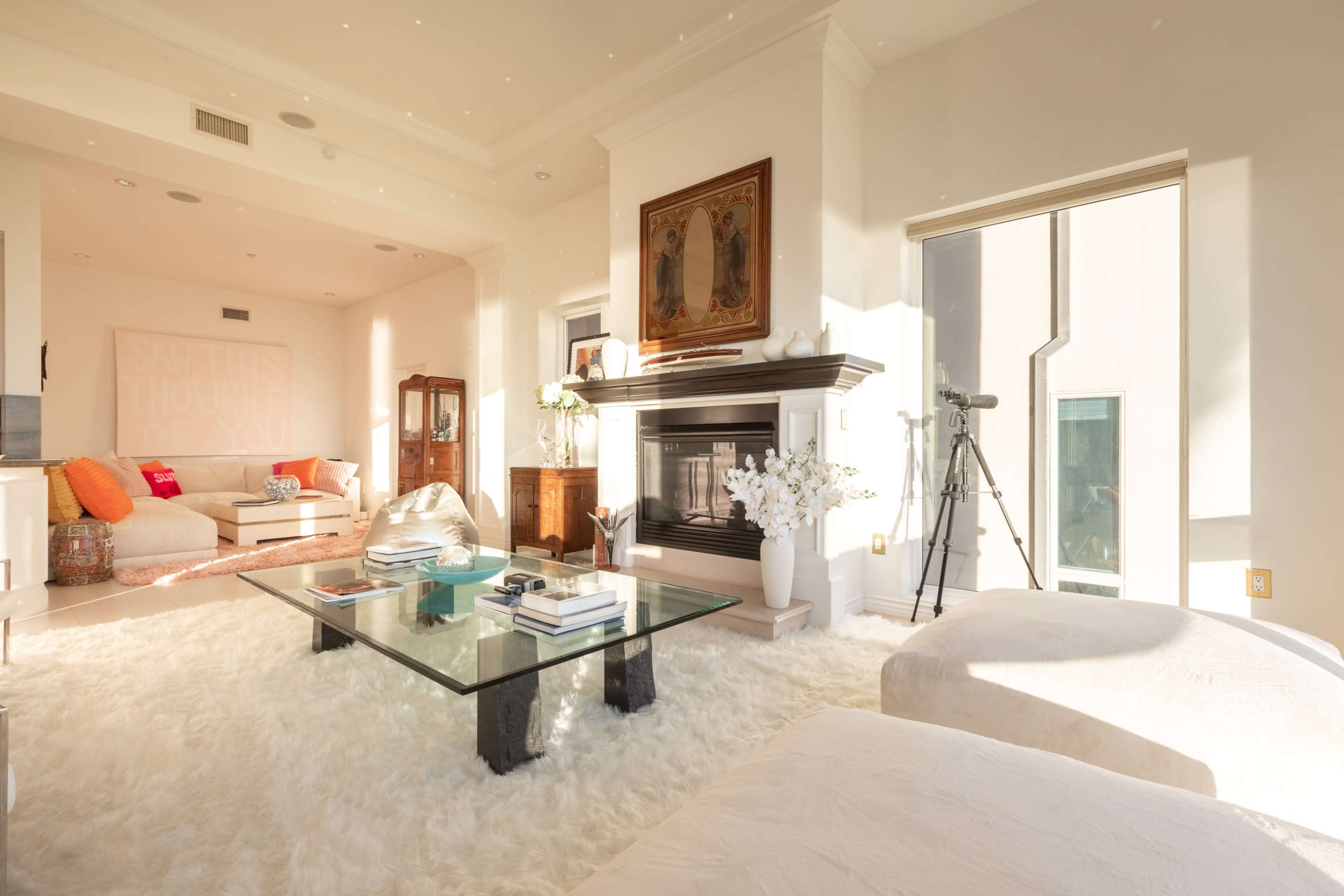 The image shows a bright, airy living room with a large window, a cozy seating area, a glass coffee table, and a fireplace.