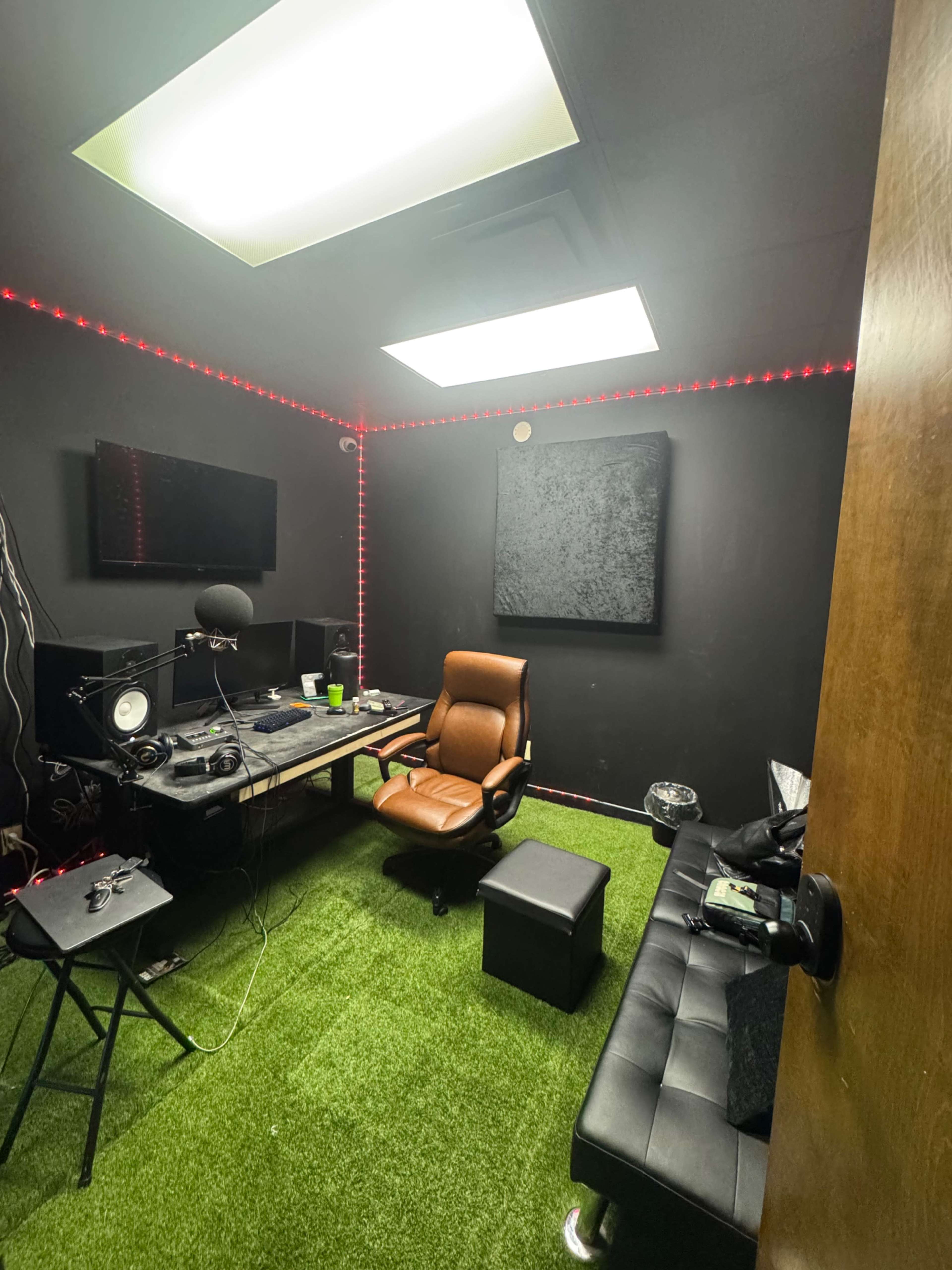 The image shows a small recording studio with a black wall, a desk with audio equipment, a brown leather chair, and artificial grass covering the floor.
