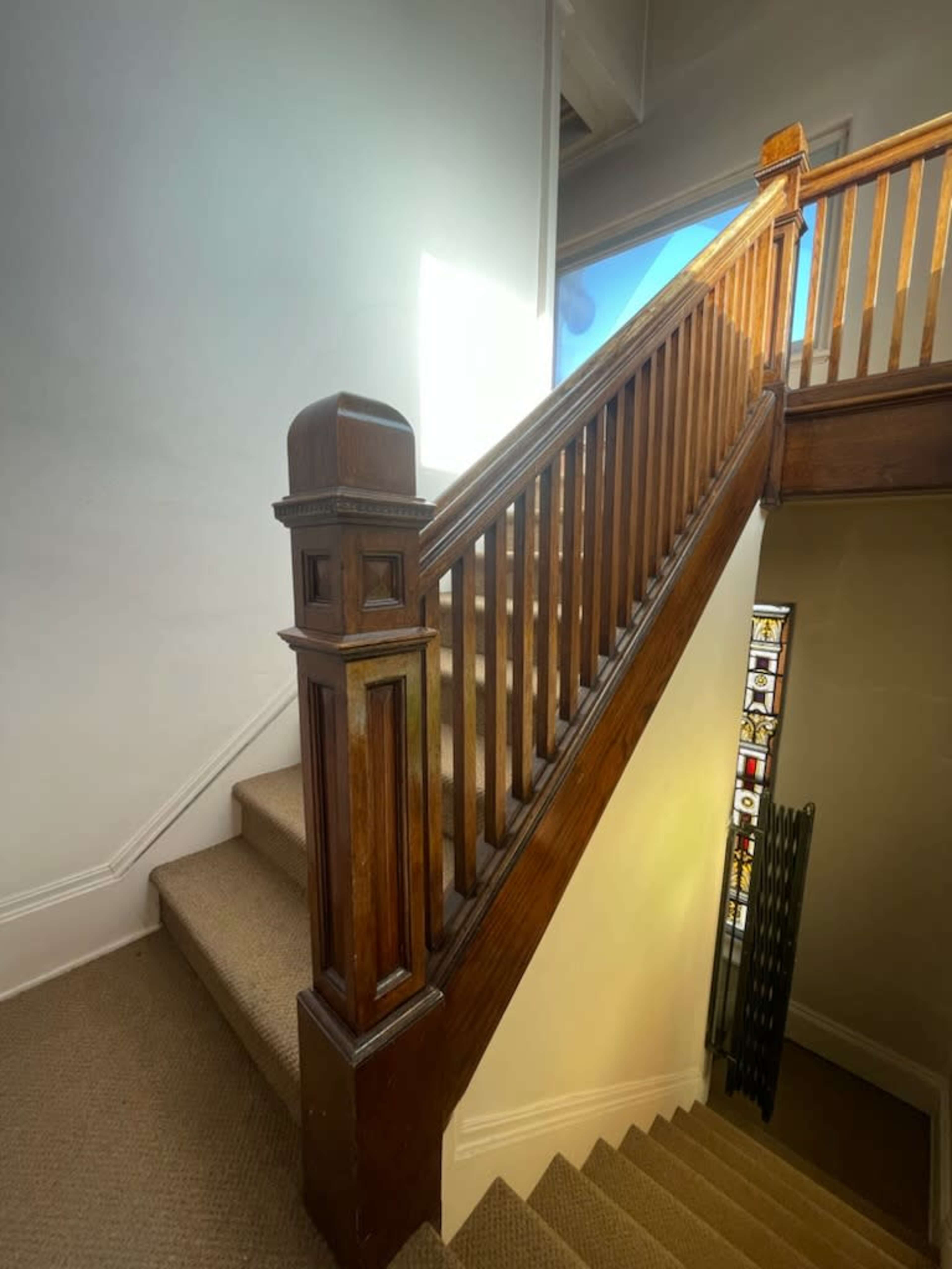 A wooden staircase with a railing leads upward to a landing near a window, accompanied by a decorative stained glass panel.