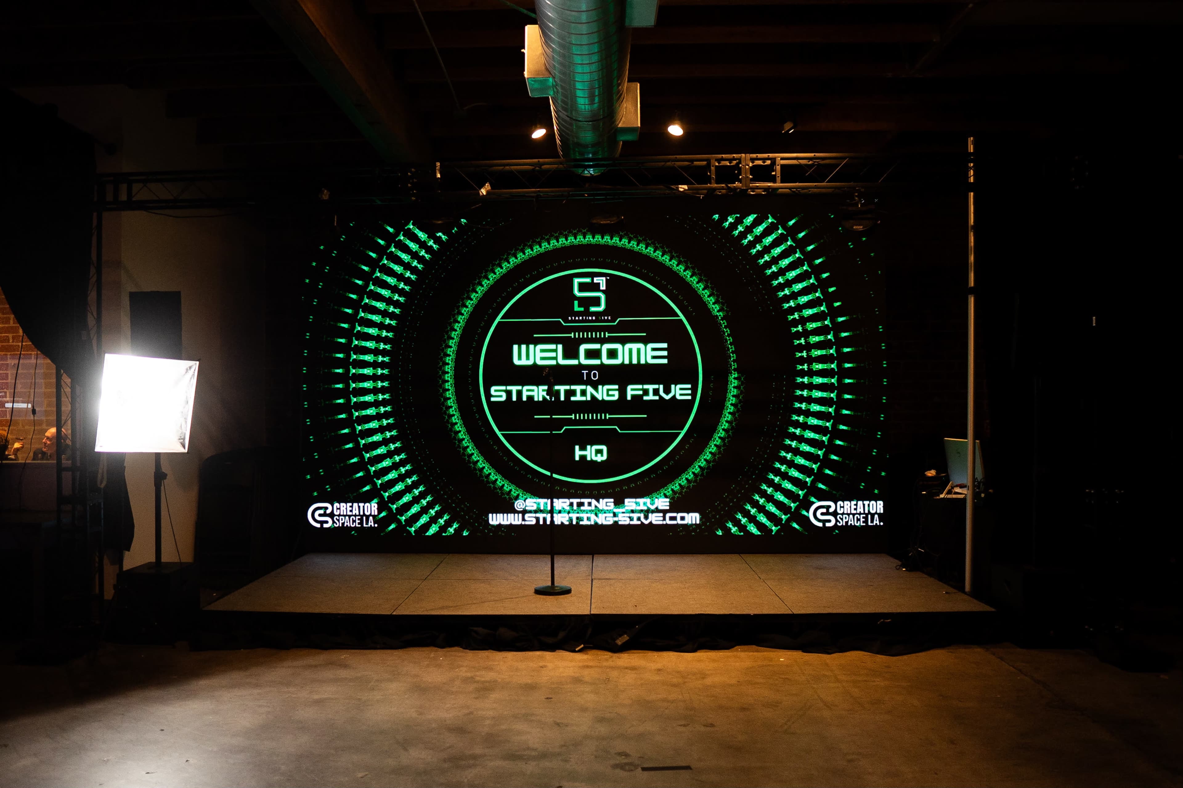 The image shows a stage setup with a large digital screen displaying "WELCOME TO STARTING FIVE" in a dimly lit event space.