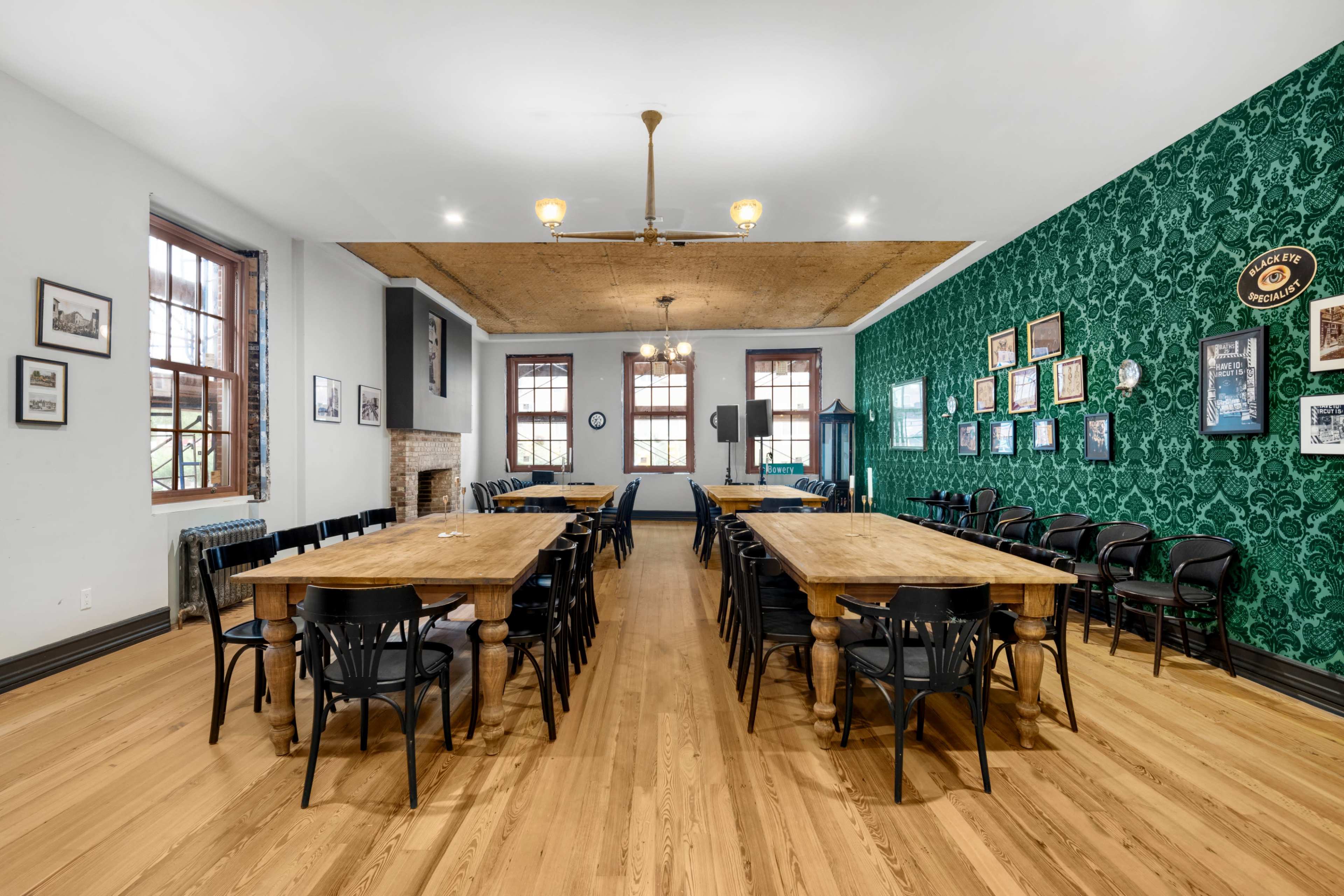 A spacious room with wooden tables arranged in a long formation, black chairs surrounding them, and green patterned wallpaper on one wall.