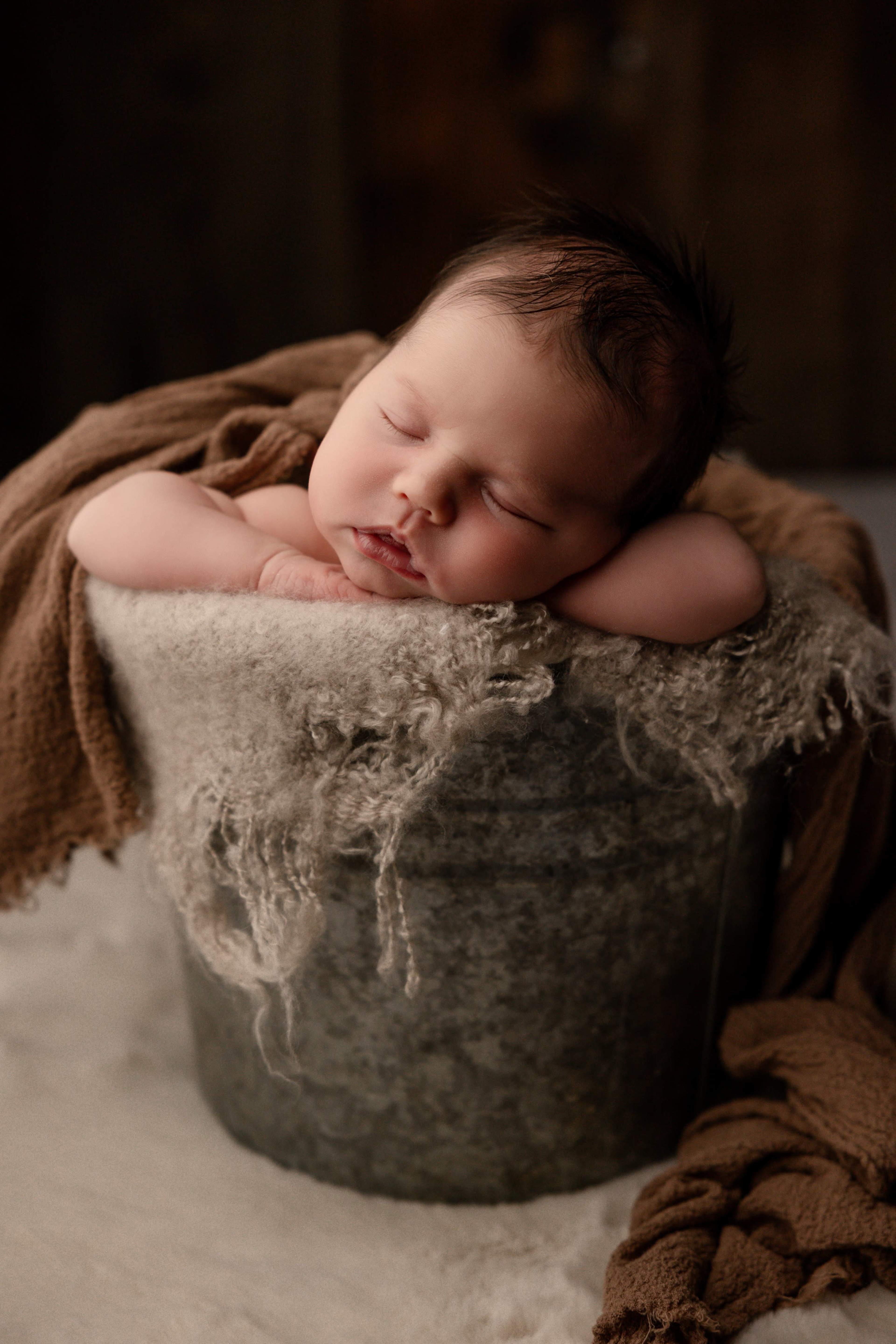 A sleeping newborn rests with their head on a rustic metal bucket draped with a soft cloth.