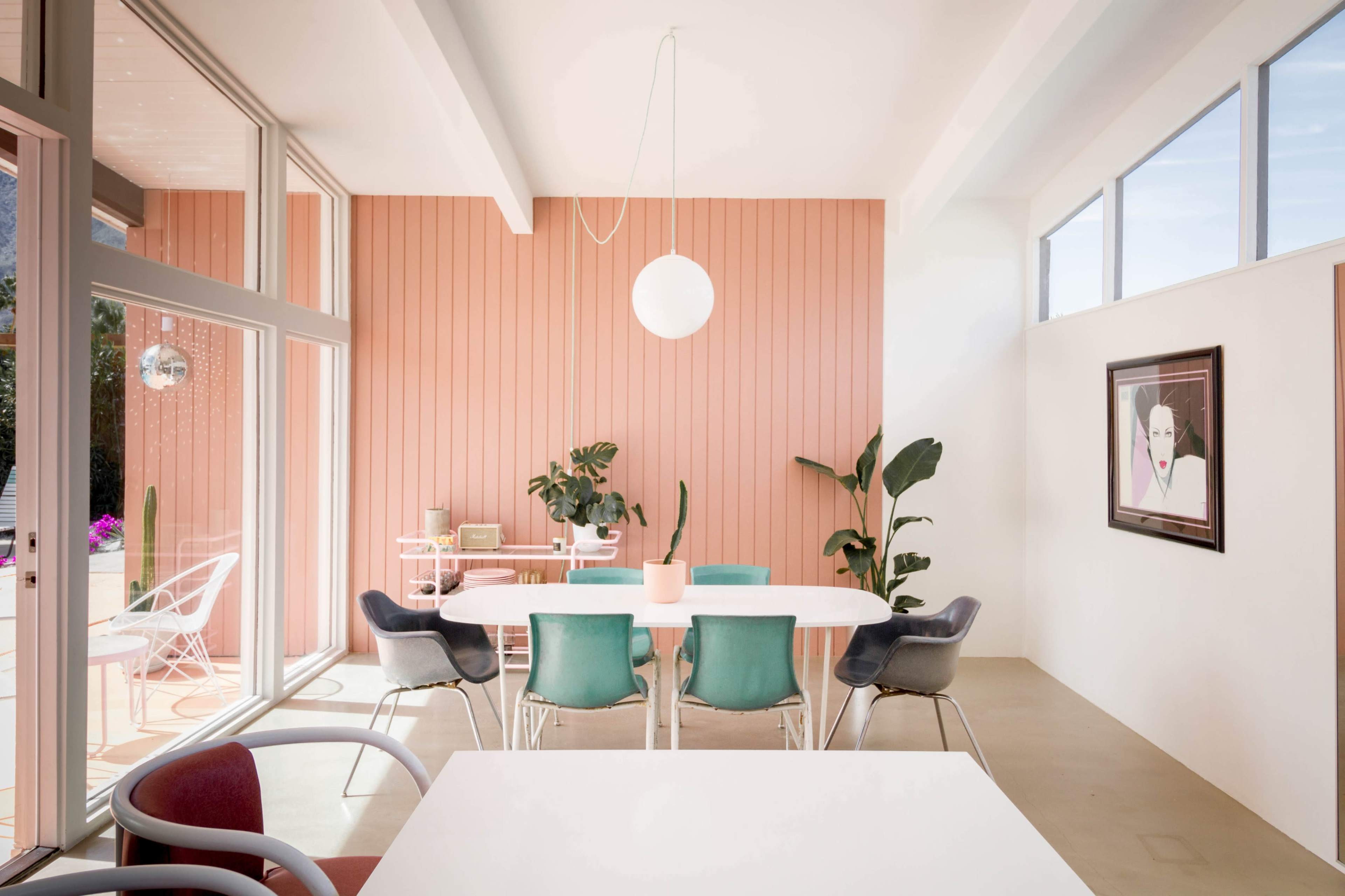A modern dining area featuring a white table surrounded by colorful chairs, large windows, and a pink wooden accent wall.