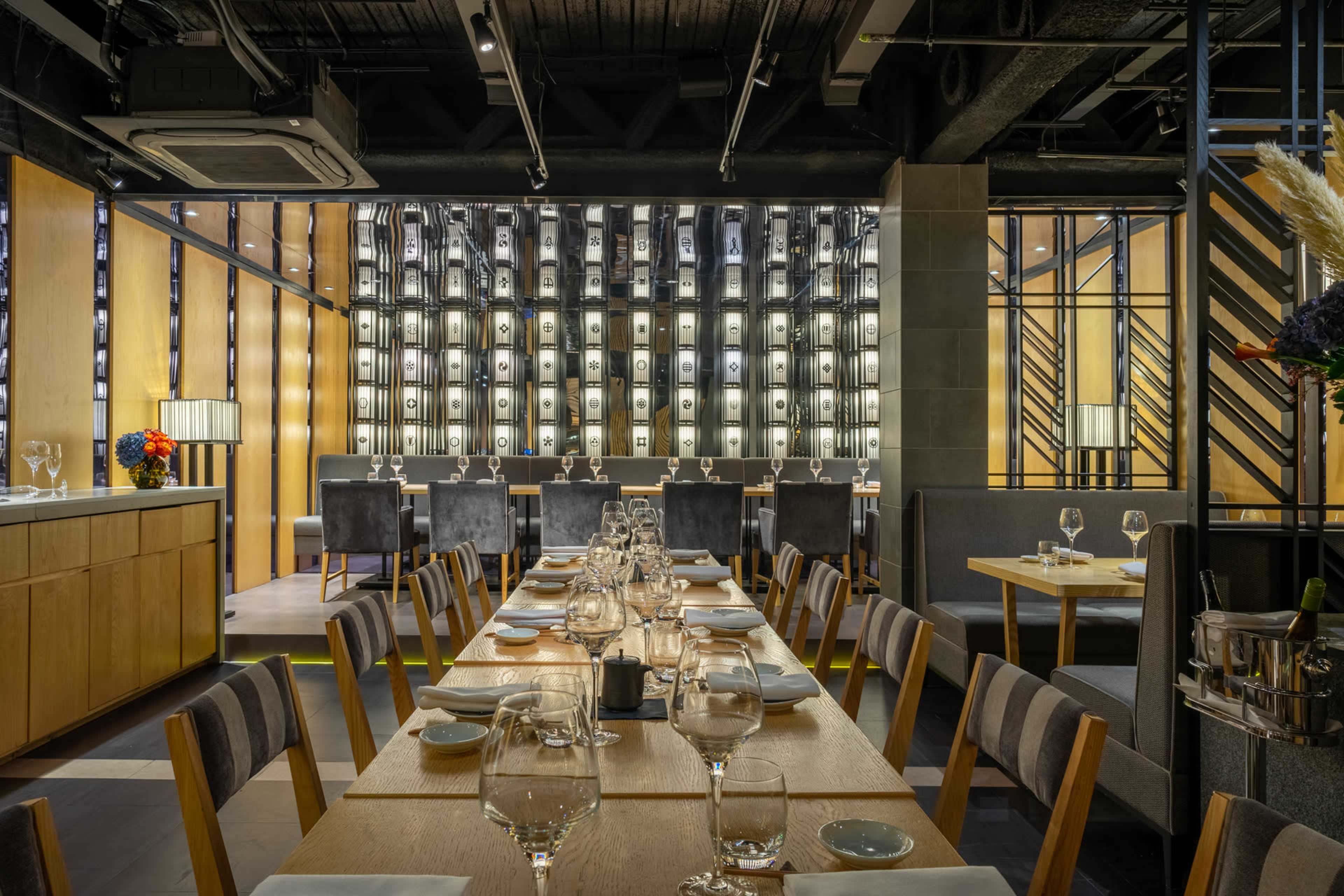 The image shows a modern restaurant interior featuring a long table set for dining, with a backdrop of illuminated wine shelves and contemporary decor.