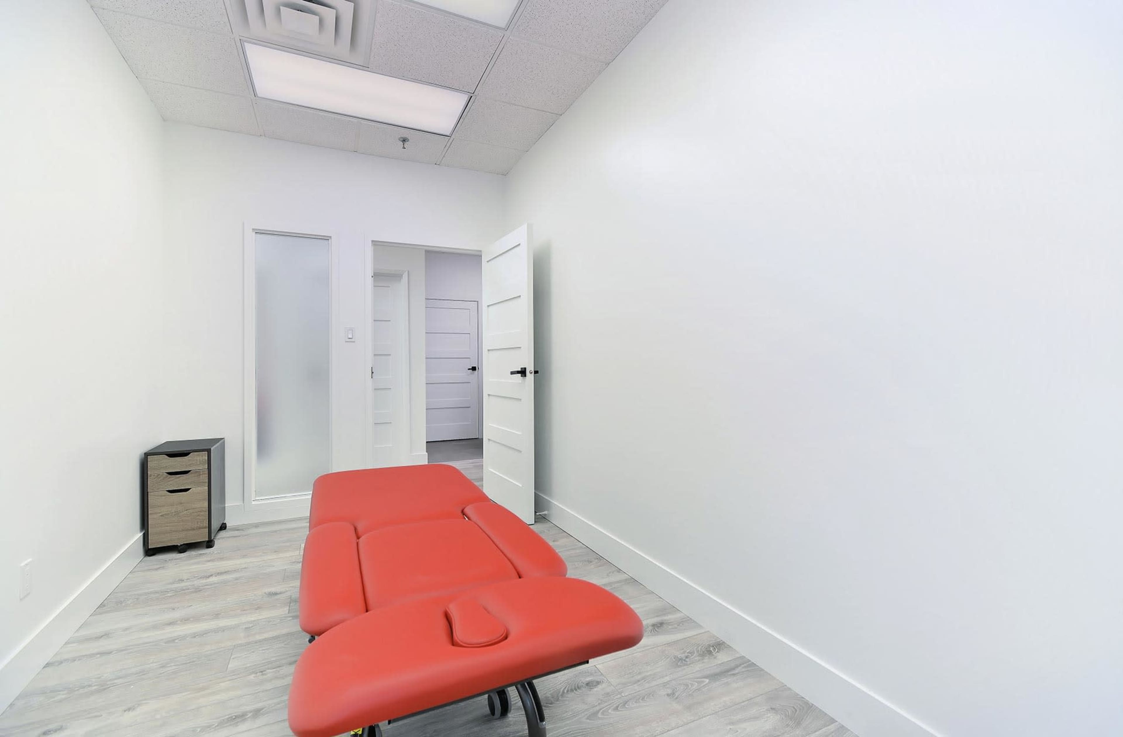 The image shows a minimalist treatment room with a red examination table and a small storage unit against a white wall.