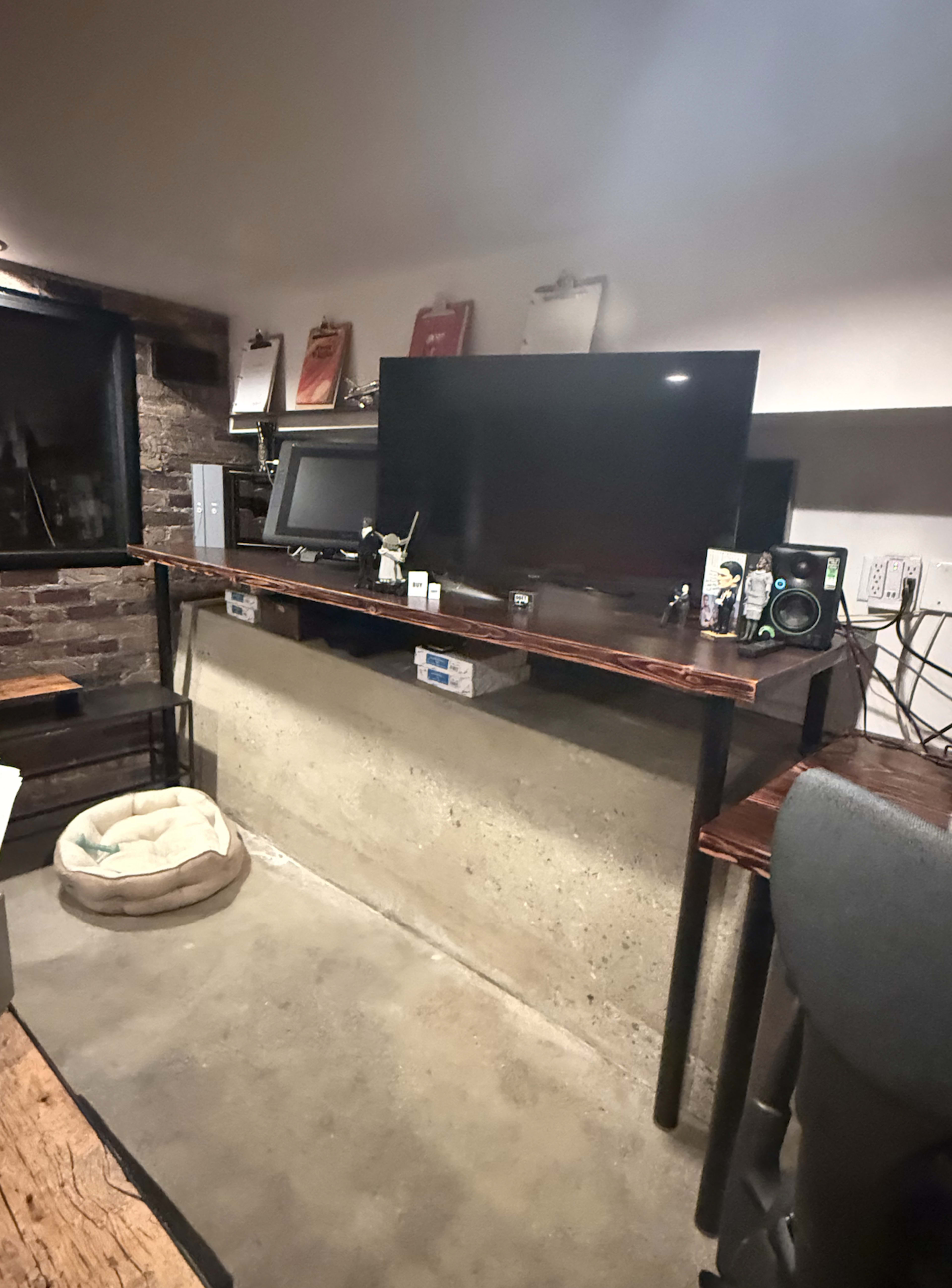 The image depicts a small workspace featuring a large monitor mounted on a wooden shelf, brick walls, and a concrete floor.