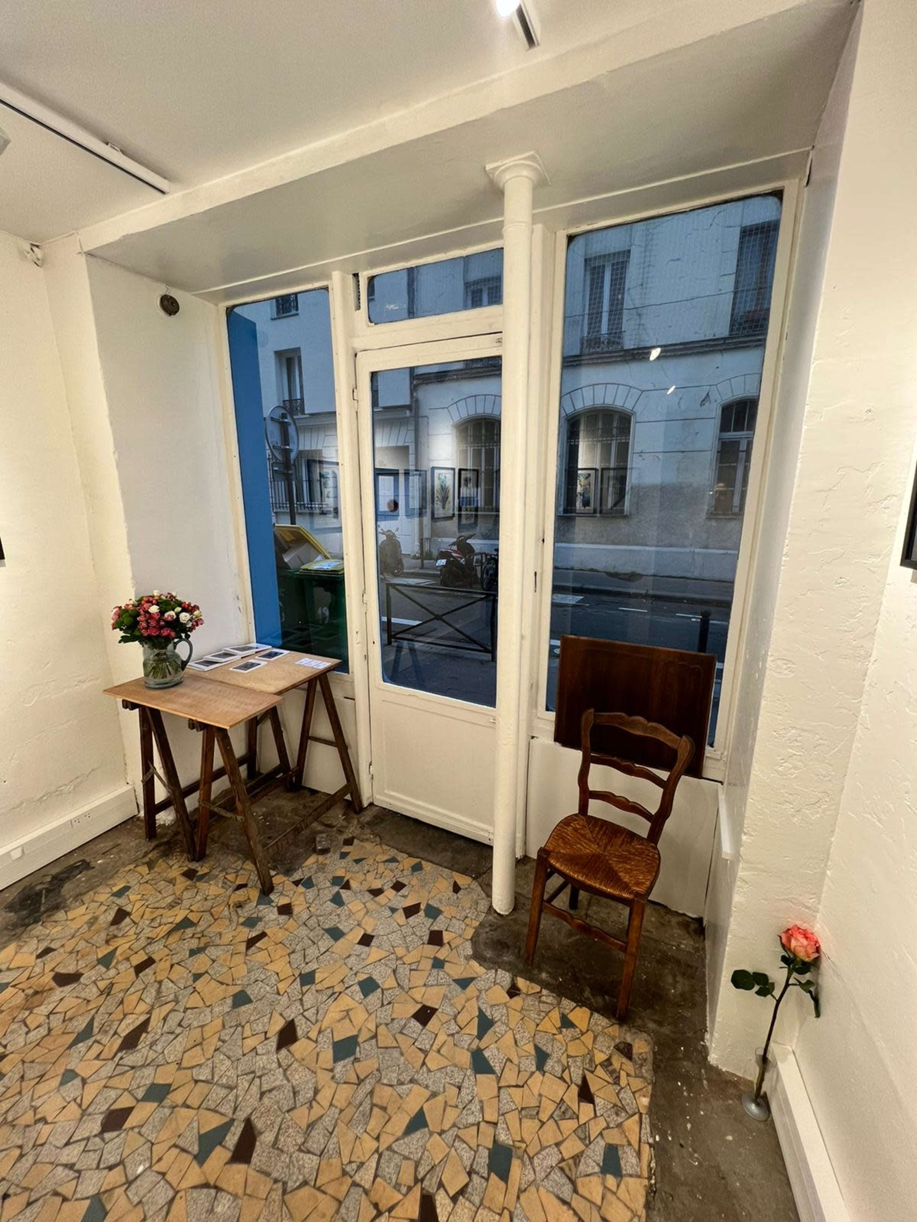 The image shows a small art gallery with a table and a wooden chair positioned near a large window overlooking a street.