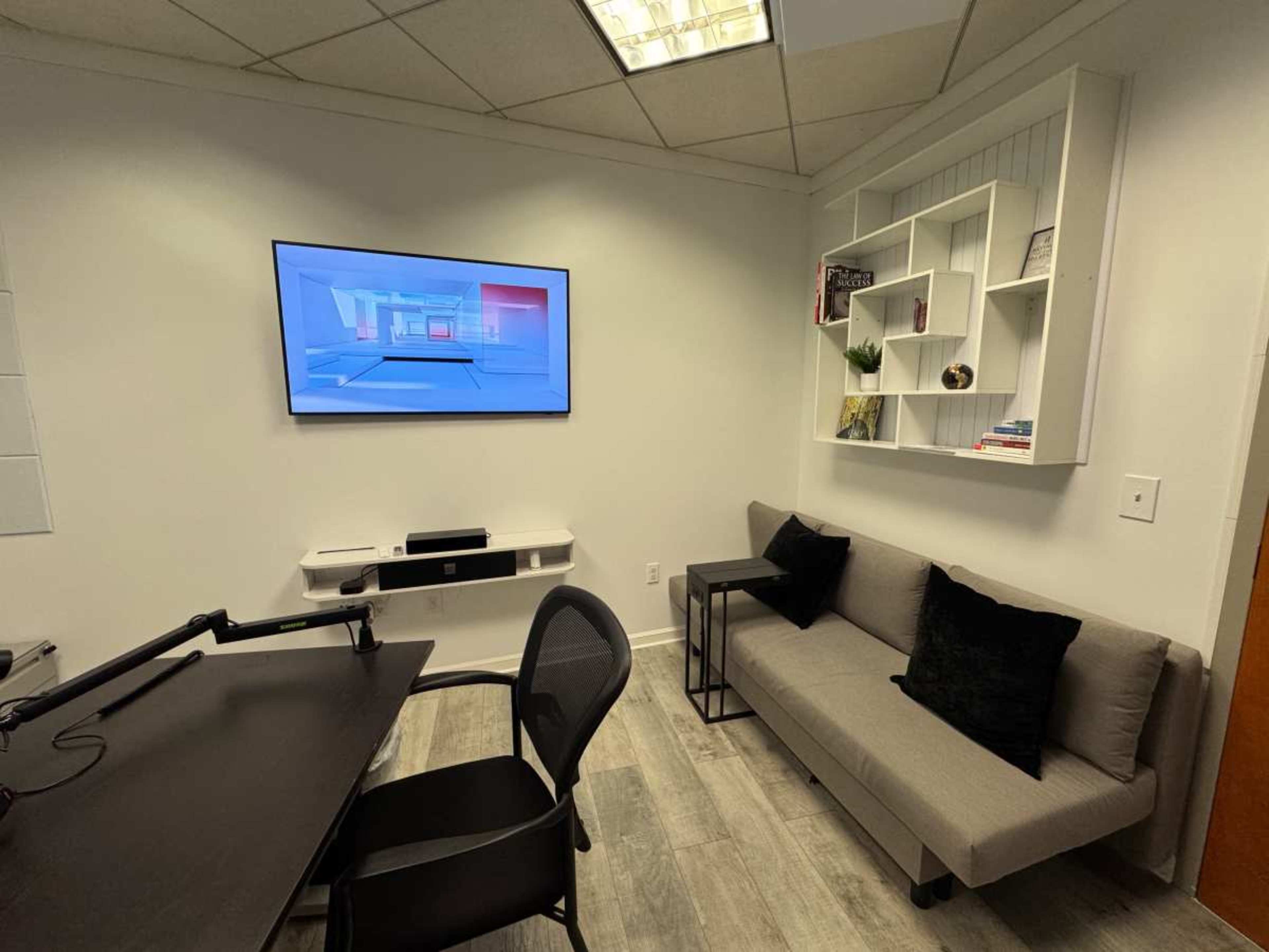 The image shows a modern office space featuring a gray sofa, a wall-mounted television, a small table, and a shelf with decorative items.