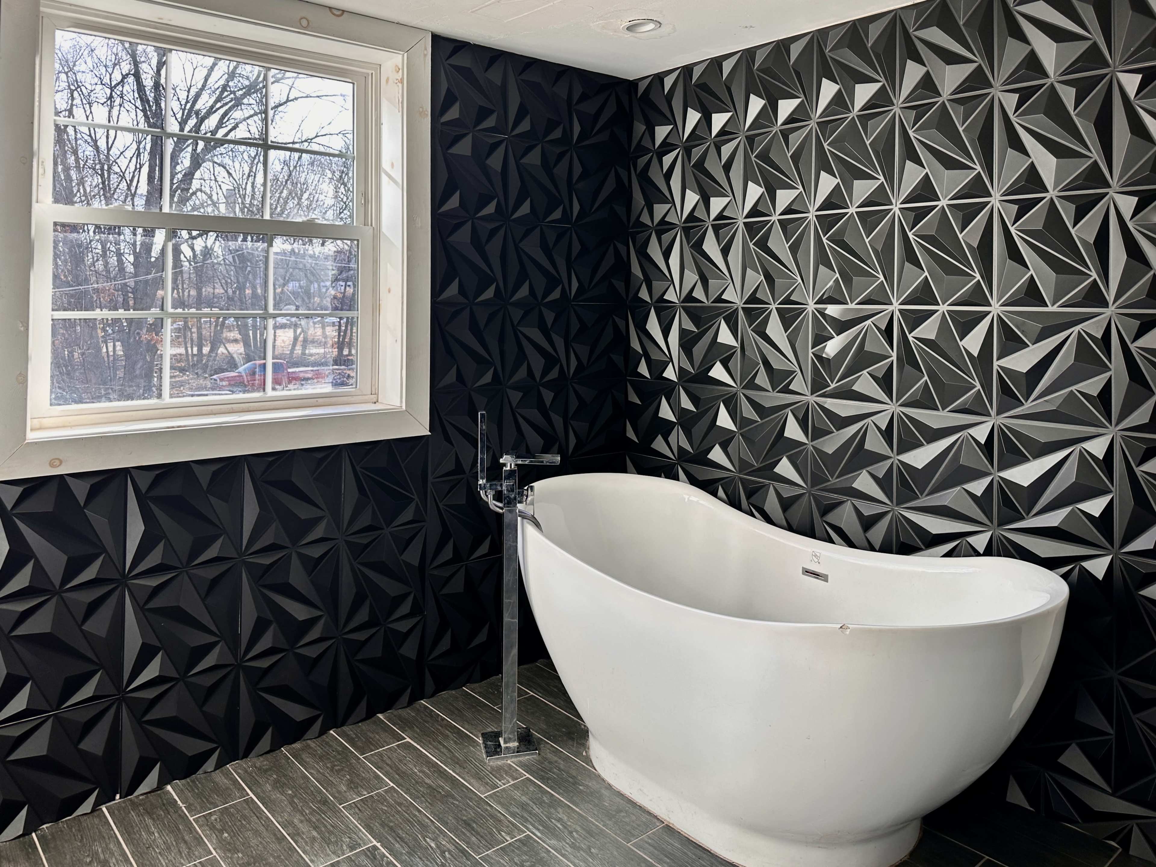 A modern bathroom featuring a white freestanding bathtub against a black geometric patterned wall and a window with trees visible outside.