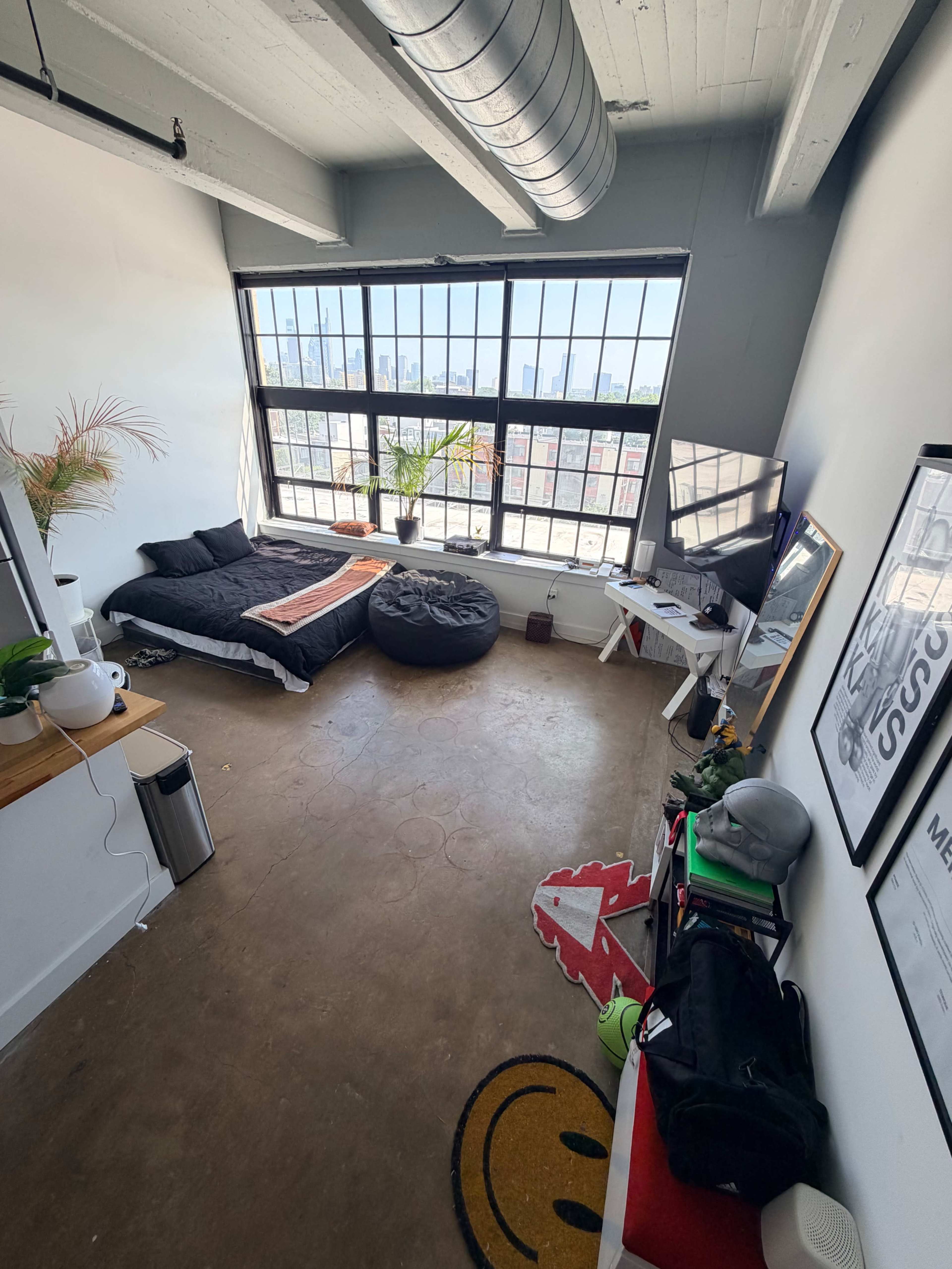 The image shows a modern bedroom with a large window overlooking a city skyline, featuring a bed, a bean bag chair, a desk, and various decorations.