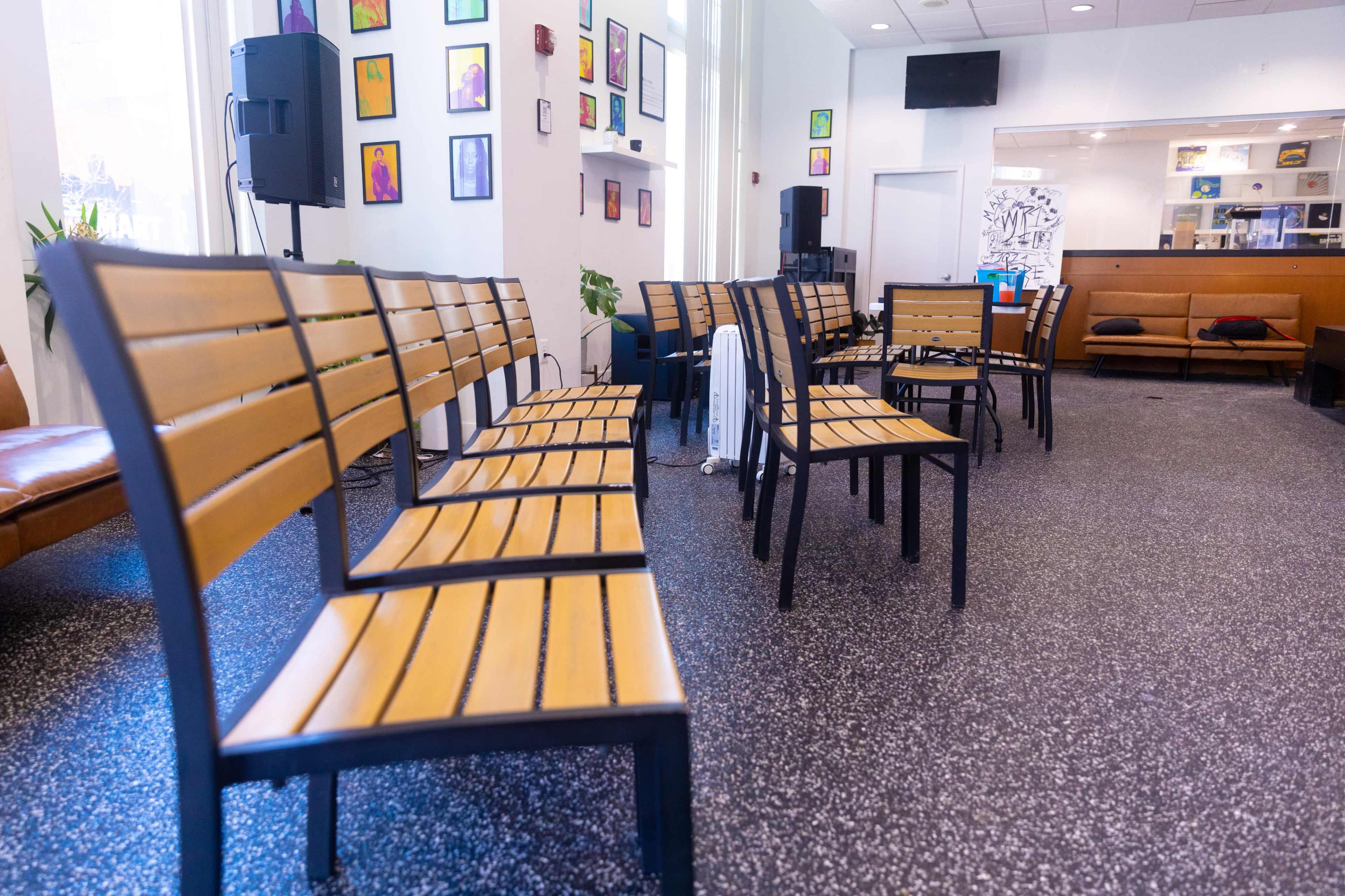 The image shows a row of wooden benches arranged in a seating area, with a small stage and colorful artwork on the walls in the background.