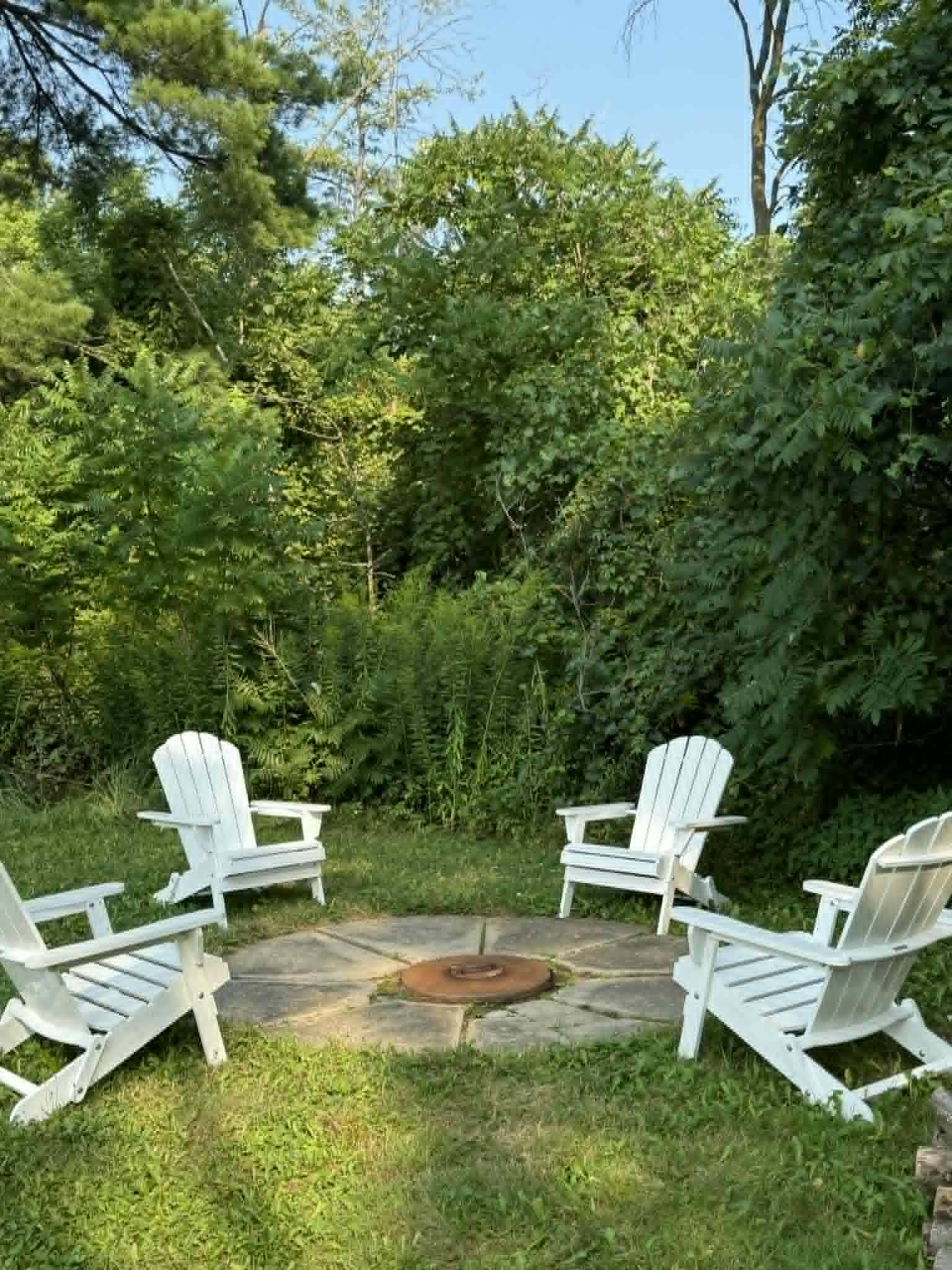 Four white Adirondack chairs are arranged in a semicircle around a stone fire pit in a green, wooded area.