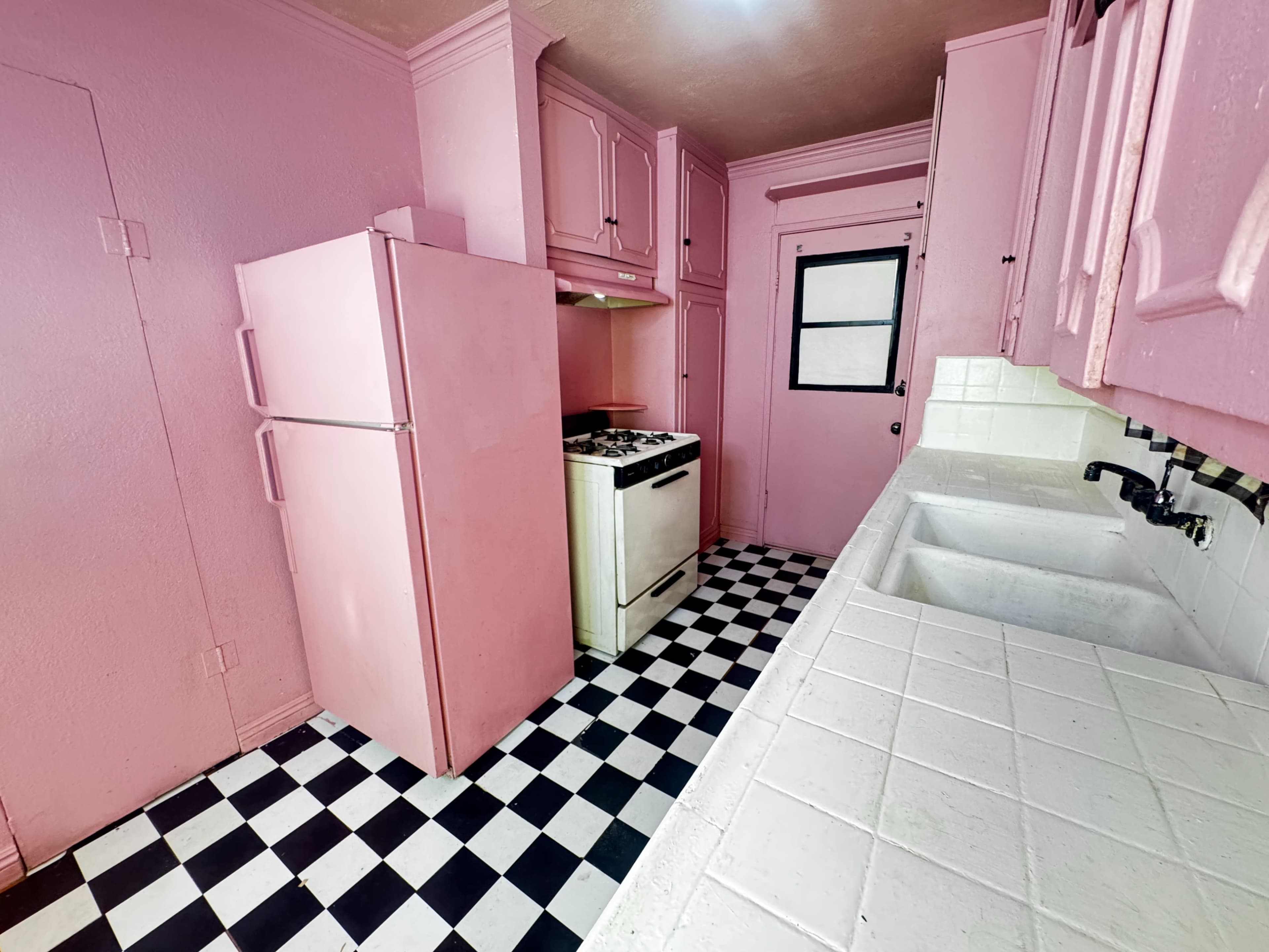 The image shows a pink kitchen with a checkerboard black and white floor, featuring a refrigerator, stove, and double sink.